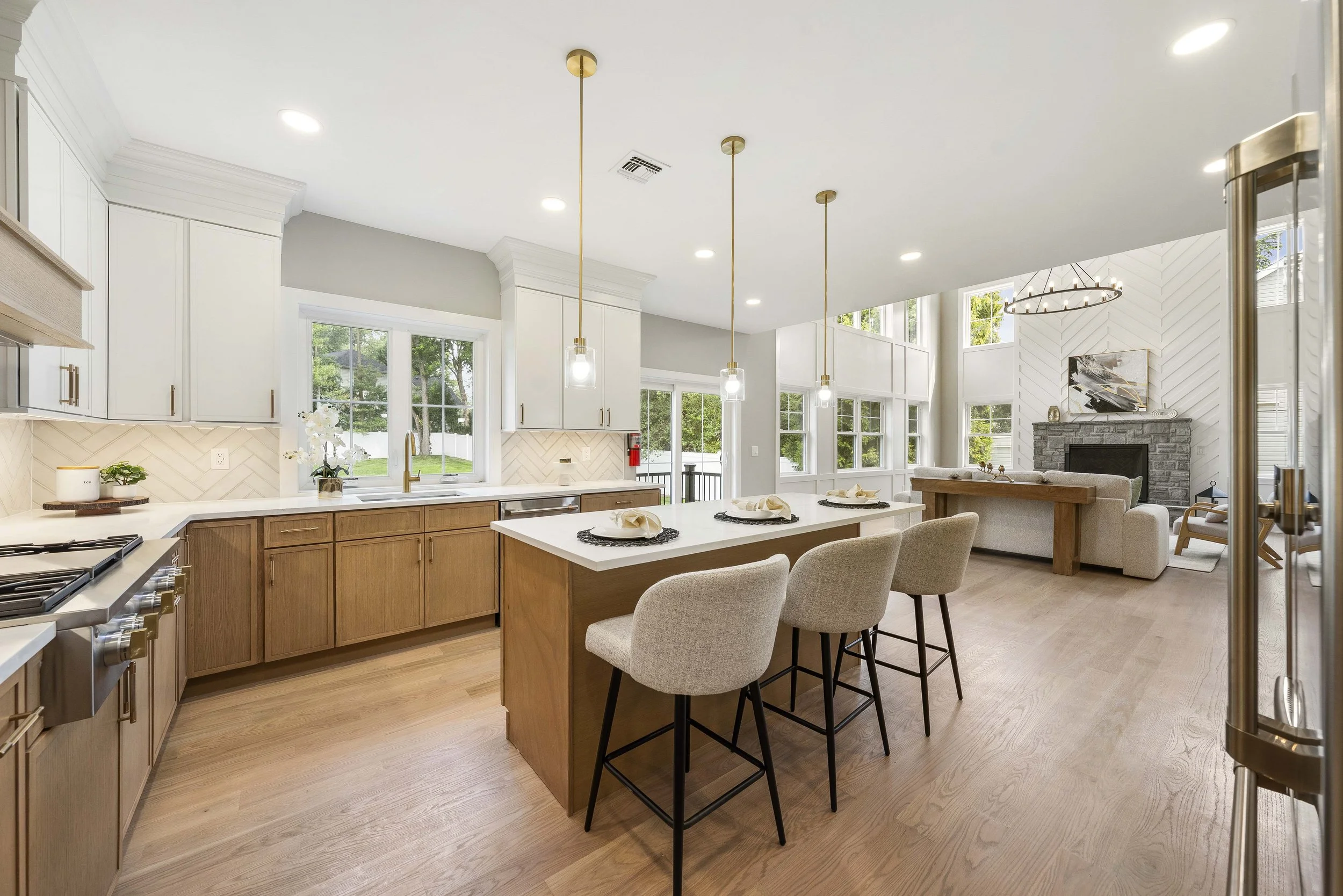 Bright open-concept kitchen and living room with white cabinets, wooden accents, pendant lighting, large windows, and a stone fireplace.