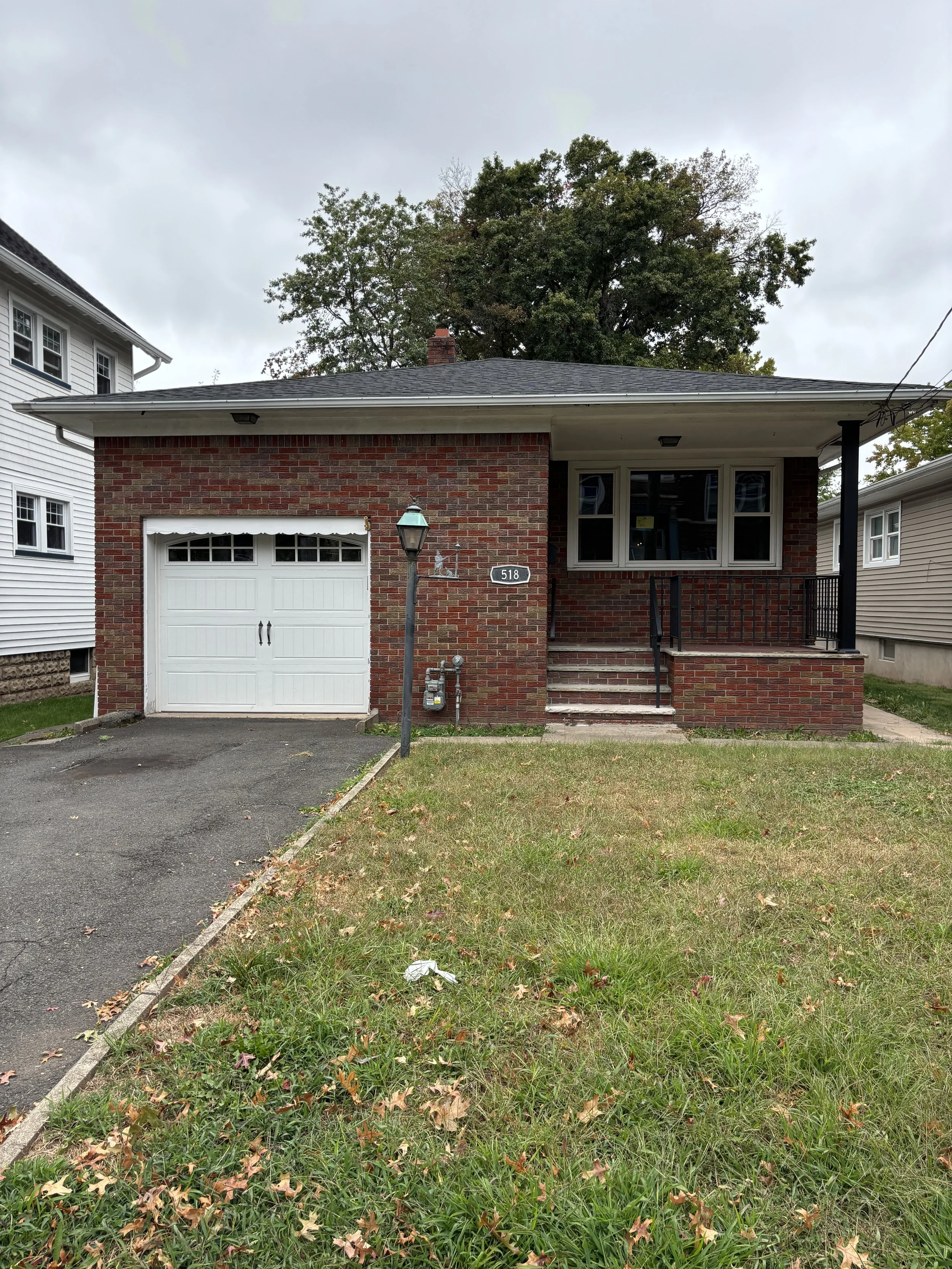 A house with a brick facade and a front porch, featuring a white garage door, steps leading to the porch, and a small lawn with grass and fallen leaves.