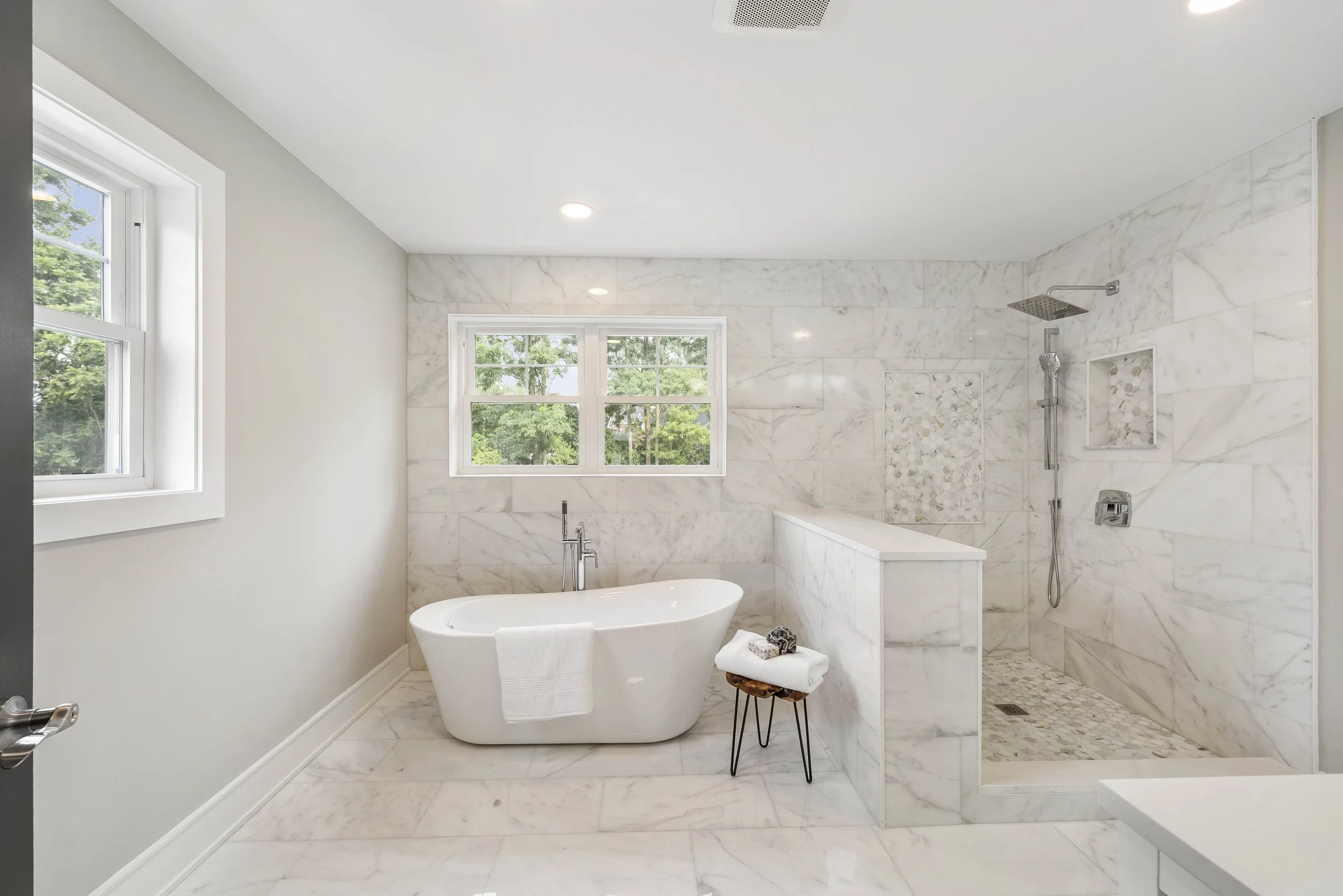 Modern bathroom with a white freestanding bathtub, marble walls, a walk-in shower with pebble floor, and large windows showing greenery outside.