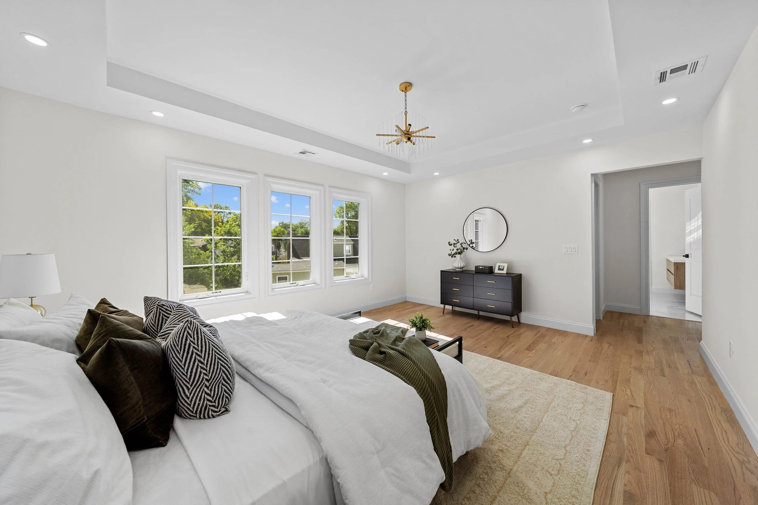 Bright bedroom with large windows, white walls, hardwood floors, a dark dresser with a mirror, and a bed with pillows and a blanket.