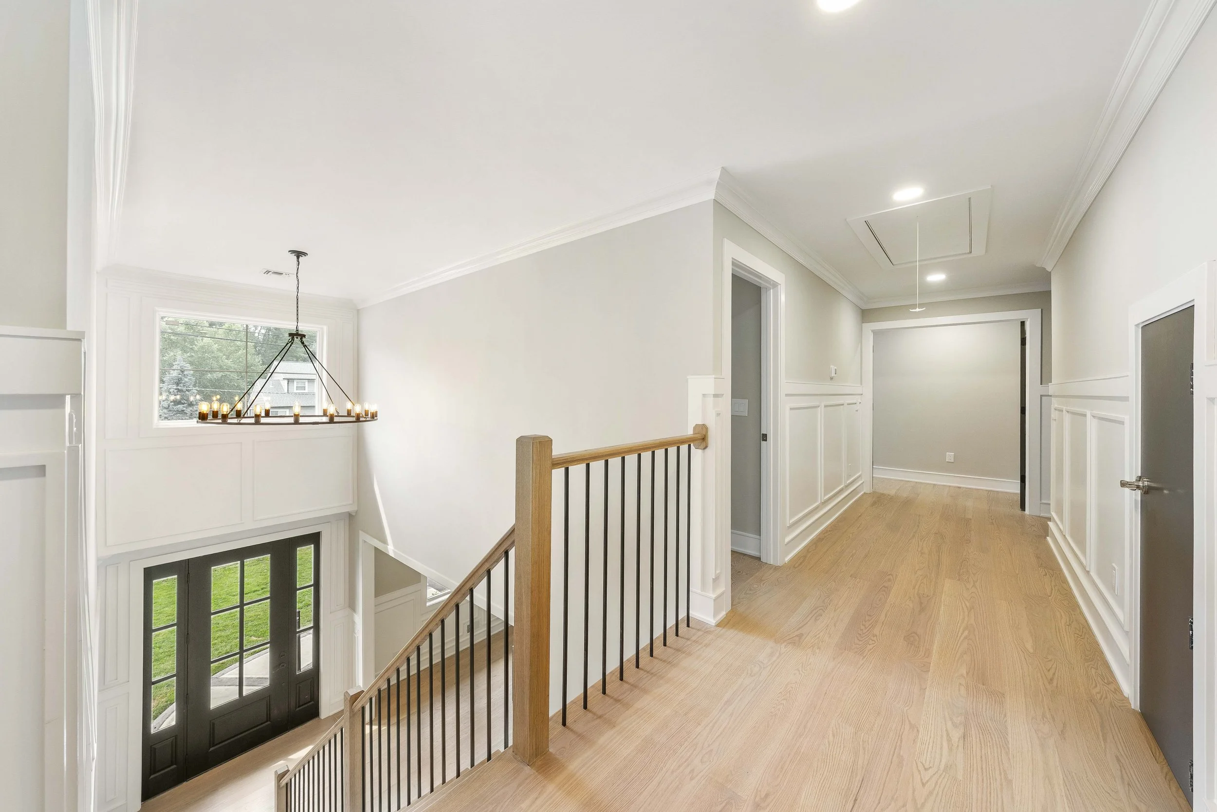 Bright and spacious upstairs hallway with white walls, wood flooring, crown molding, and a staircase with a wooden handrail leading downstairs. There are doors along the hallway and recessed ceiling lights. A window with a black chandelier hangs abov