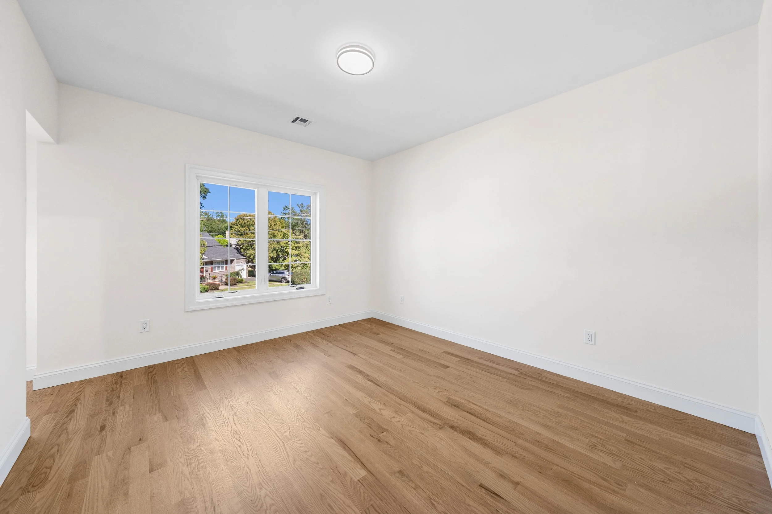 Empty room with white walls, hardwood floor, and a window showing a suburban neighborhood with trees, houses, and a parked car.