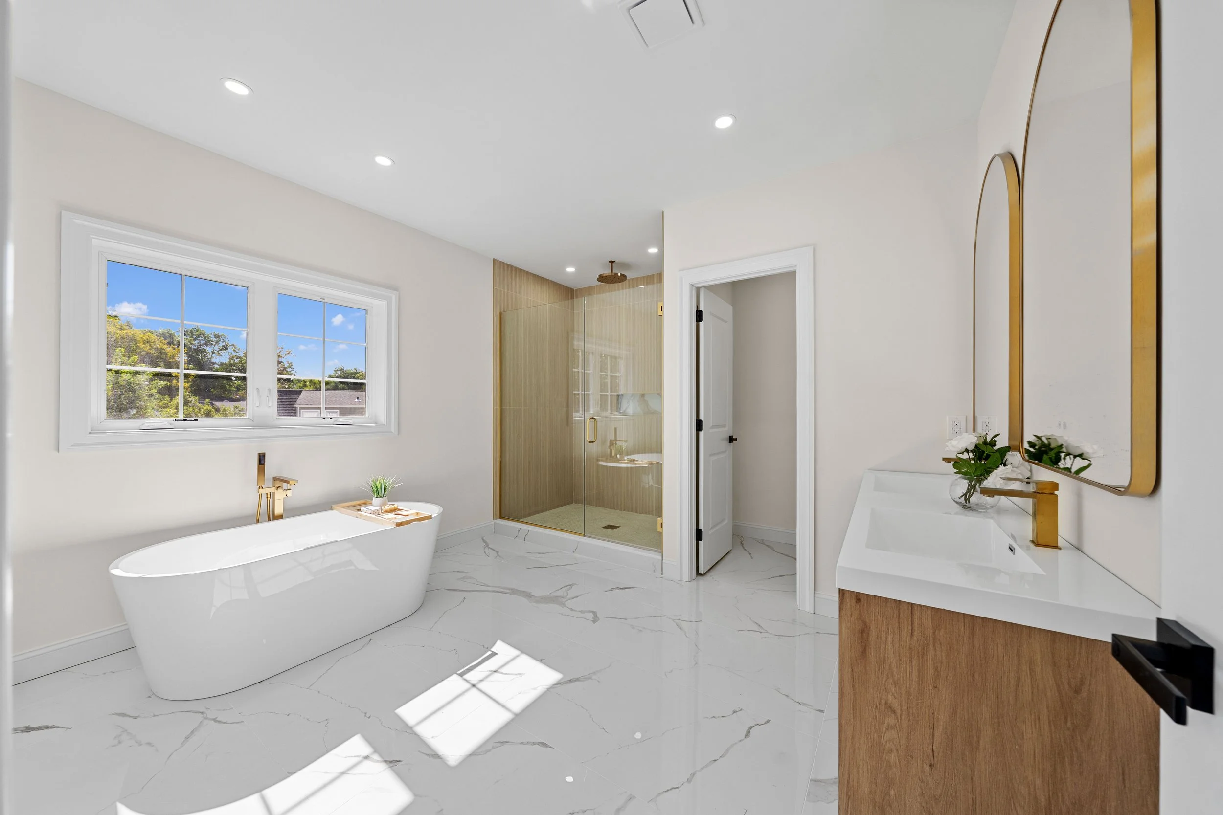 Modern bathroom with white freestanding tub, wooden accents, gold fixtures, marble floor, glass shower, white walls, and a window showing trees outside.