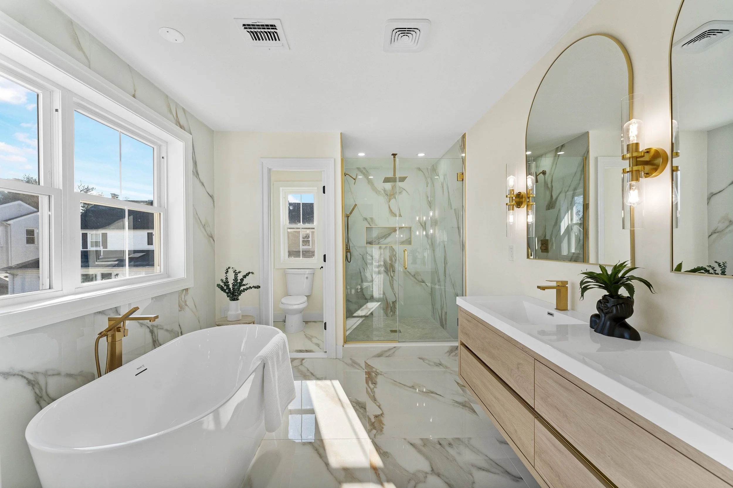 Modern bathroom featuring a freestanding bathtub with gold fixtures, marble walls and floors, a walk-in glass shower with gold accents, a wooden vanity with a large mirror, and a toilet near a window with natural light.