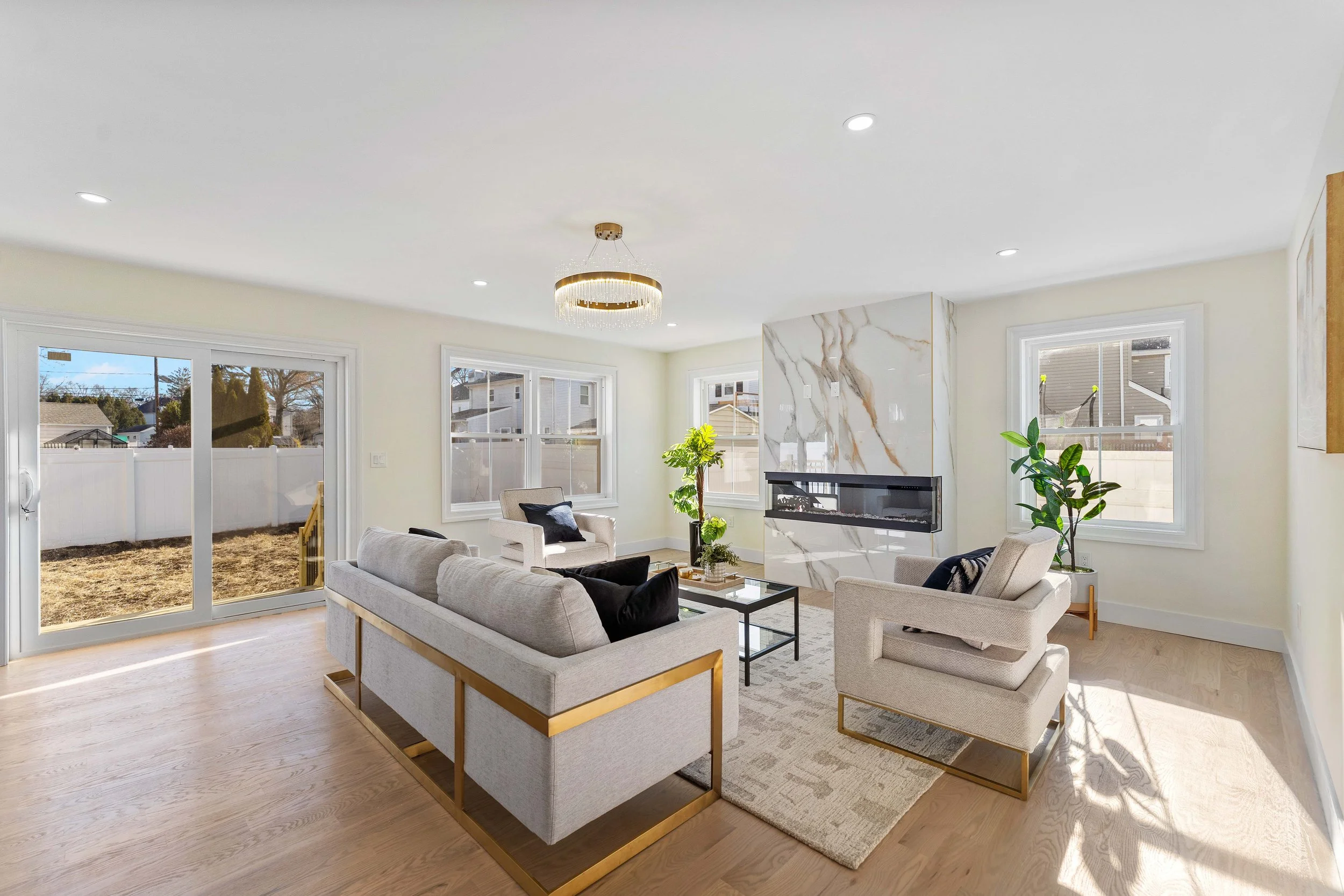 Bright living room with beige sofas, armchairs, a glass-top coffee table, a marble fireplace, large windows, and a sliding glass door overlooking a backyard with a white fence.