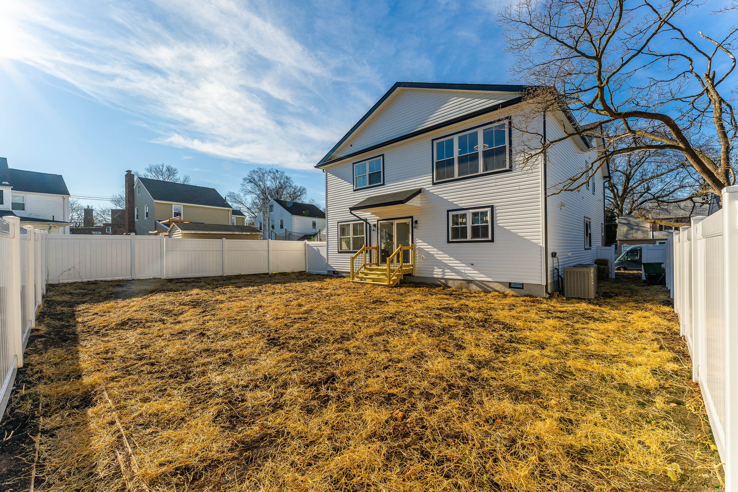 Backyard with yellowish grass, white fenced yard, white two-story house with black trim, small front porch, and a leafless tree, under a blue sky with some clouds.