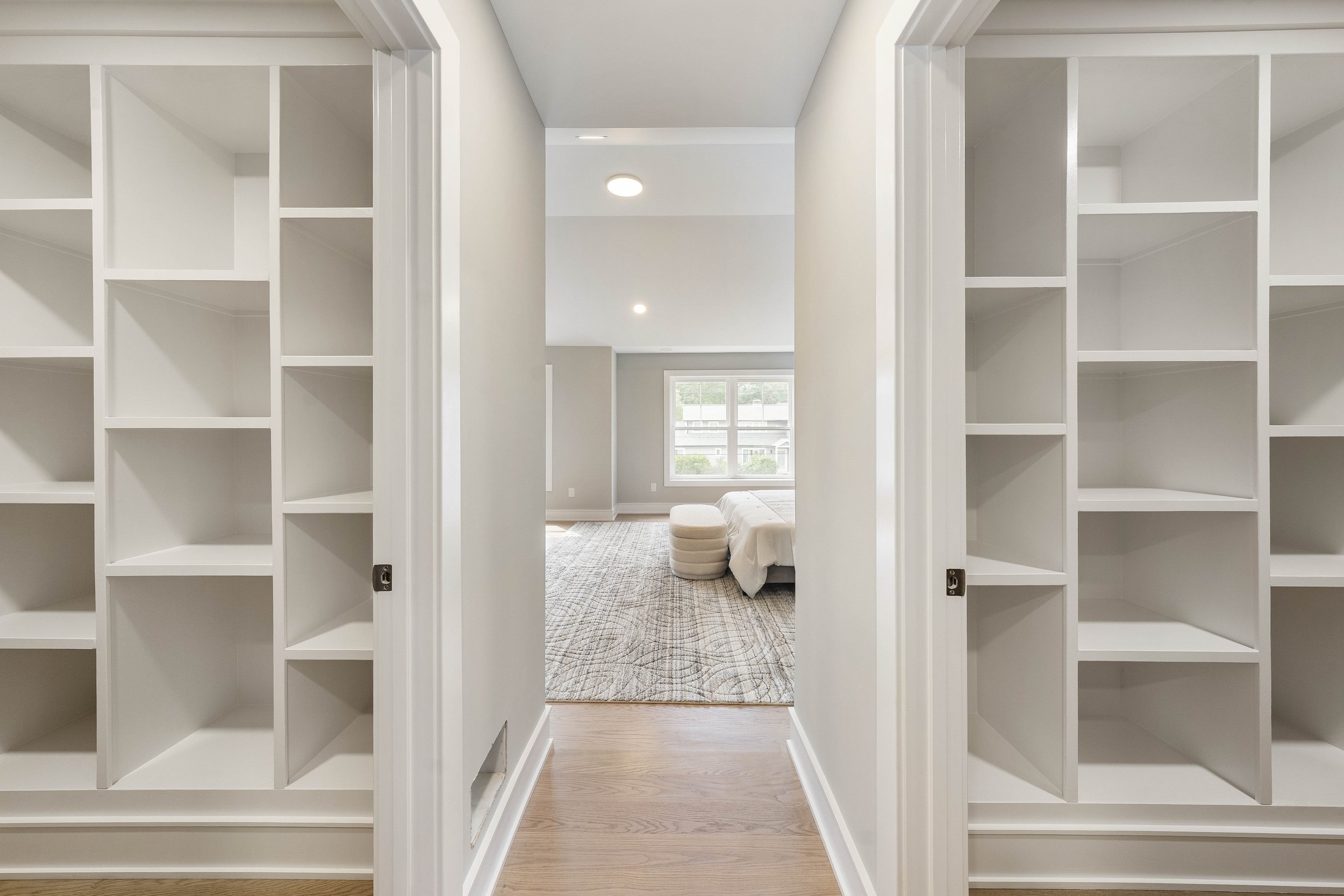 Empty built-in white bookshelves in a hallway leading to a living room with a window, ceiling lights, a sofa, and a patterned rug.