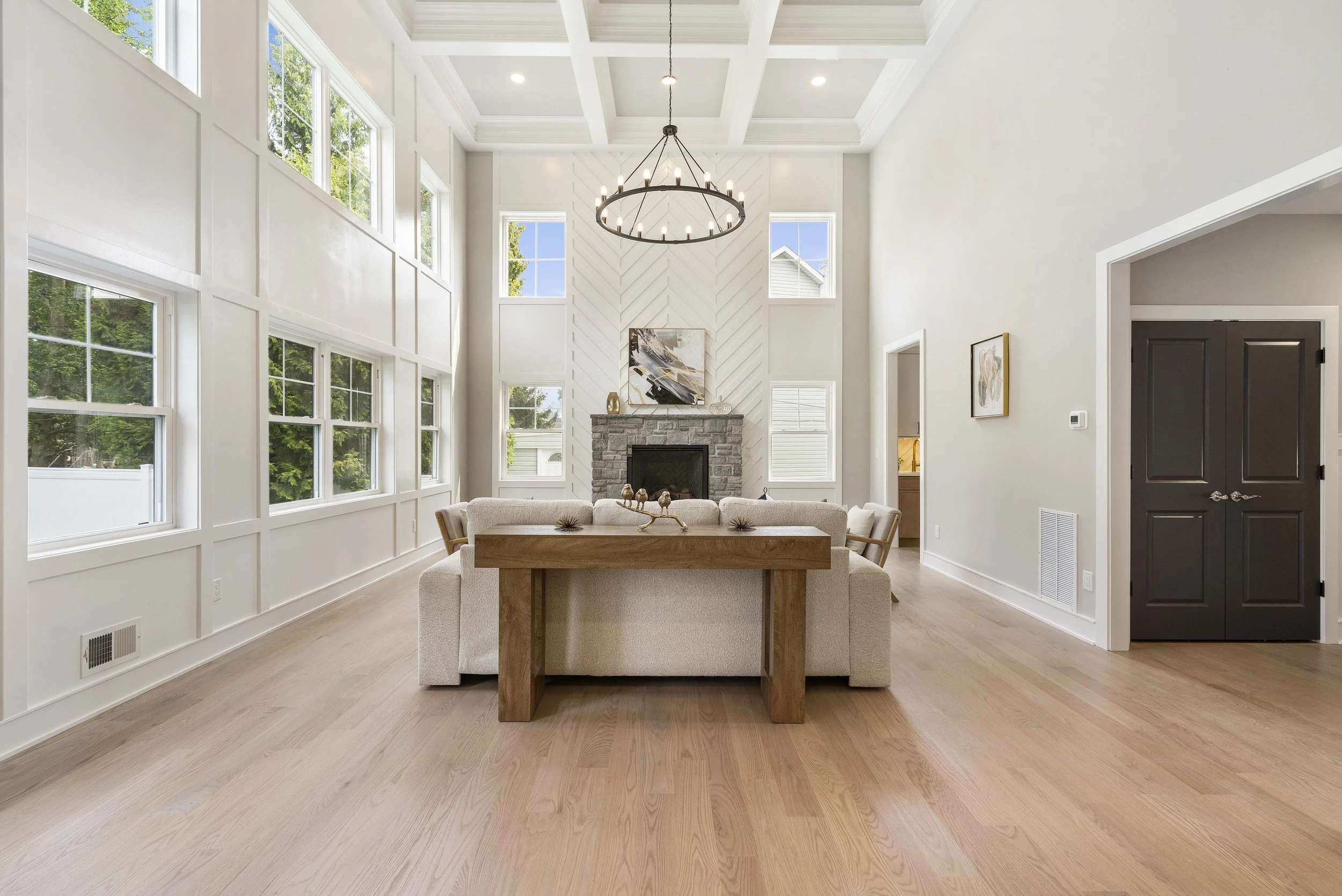 Bright, spacious living room with high ceilings, large windows, a chandelier, a stone fireplace, a light-colored sectional sofa, and a wooden dining table.