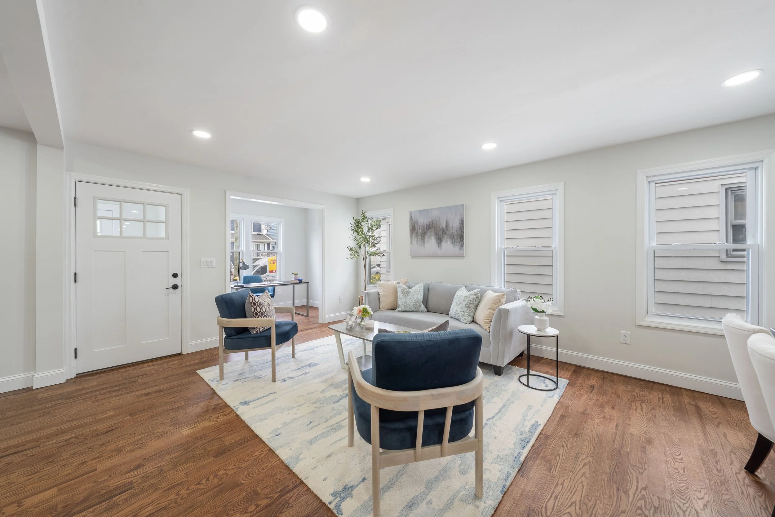 Bright living room with white walls, hardwood floors, a white sofa with decorative pillows, two navy armchairs, a small white side table with a white vase, a coffee table, and a large area rug. Windows with white trim and an artwork on the wall are visible.