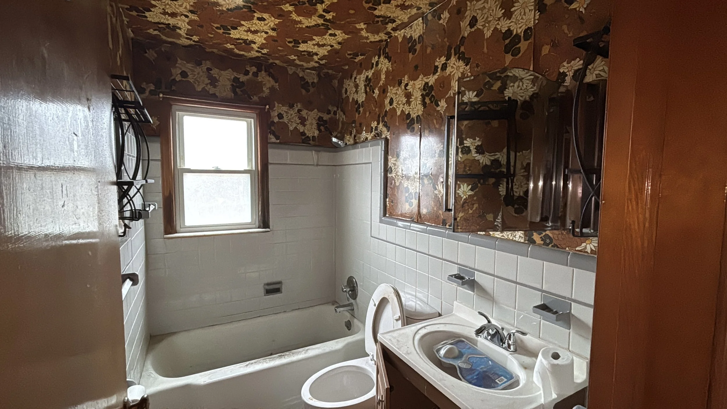 Small bathroom with vintage floral wallpaper, white tile walls, a window, bathtub, toilet, and a small sink with a mirror above.