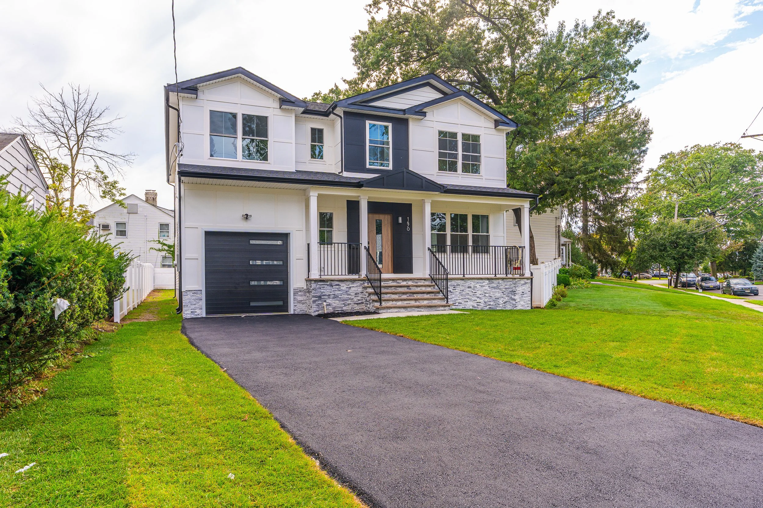A modern two-story house with white and black exterior, front porch with stairs, black driveway, green lawn, and trees in a suburban neighborhood.