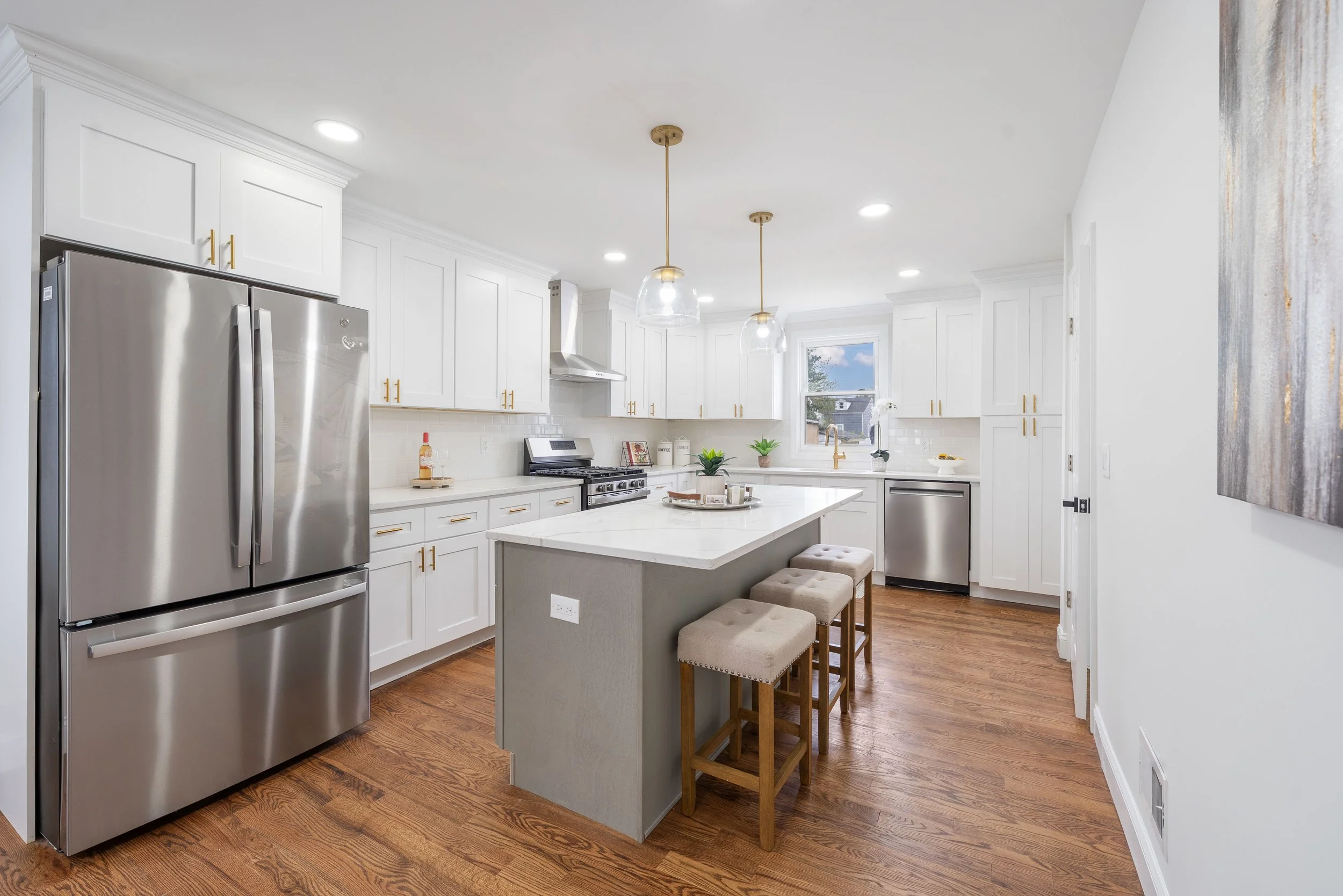 Modern kitchen with white cabinets, stainless steel appliances, a white island with a gray base, and three beige barstools. Contains hardwood floors and pendant lighting.