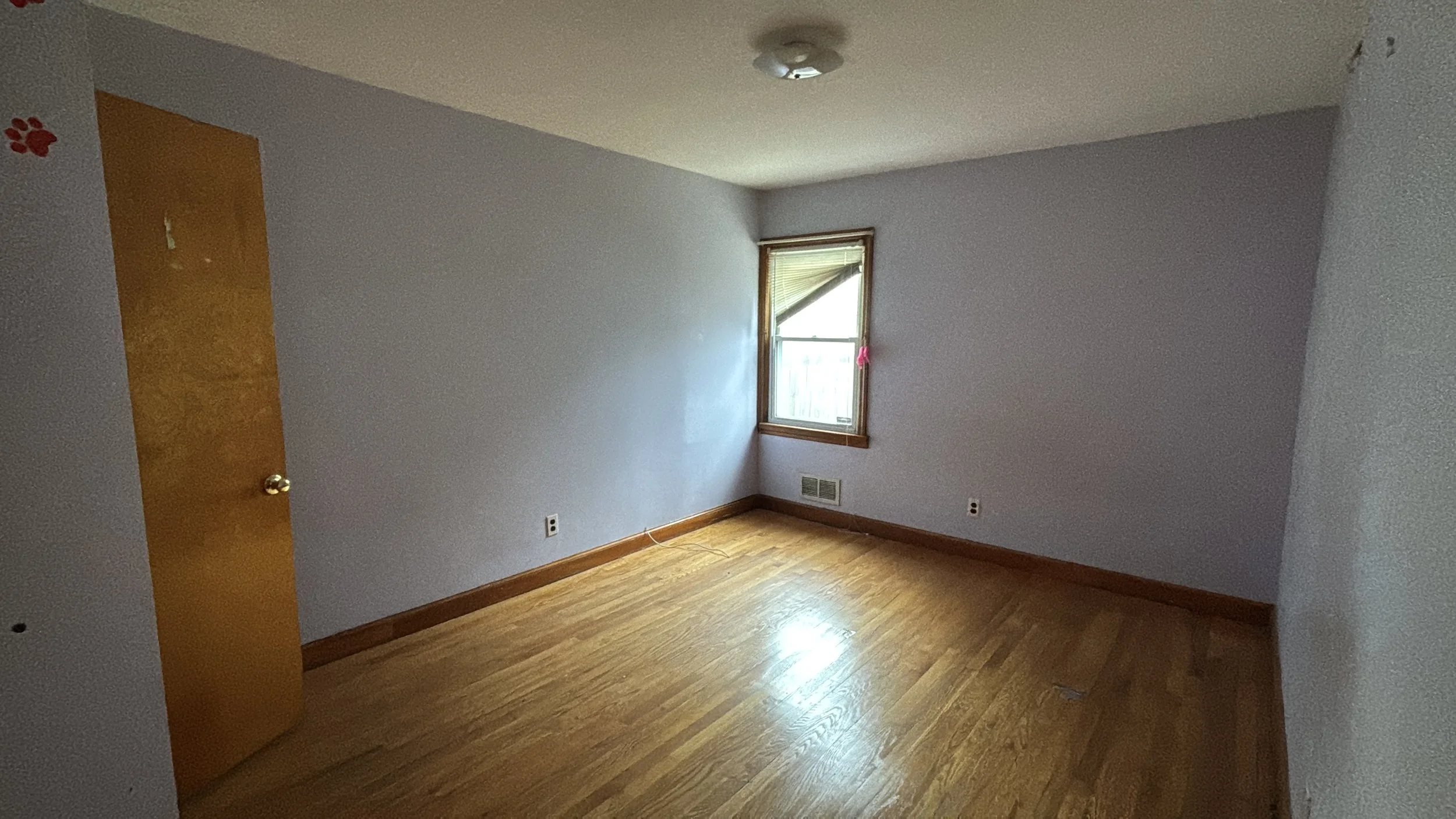 Empty room with light purple walls, hardwood floors, a small window with blinds, and a closed wooden door.