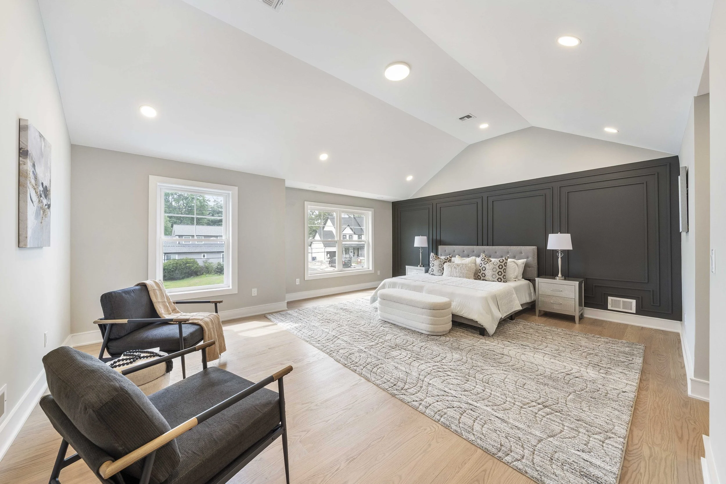A spacious, modern bedroom with a large bed, nightstands with lamps on each side, a patterned area rug, two chairs, and large windows allowing natural light.