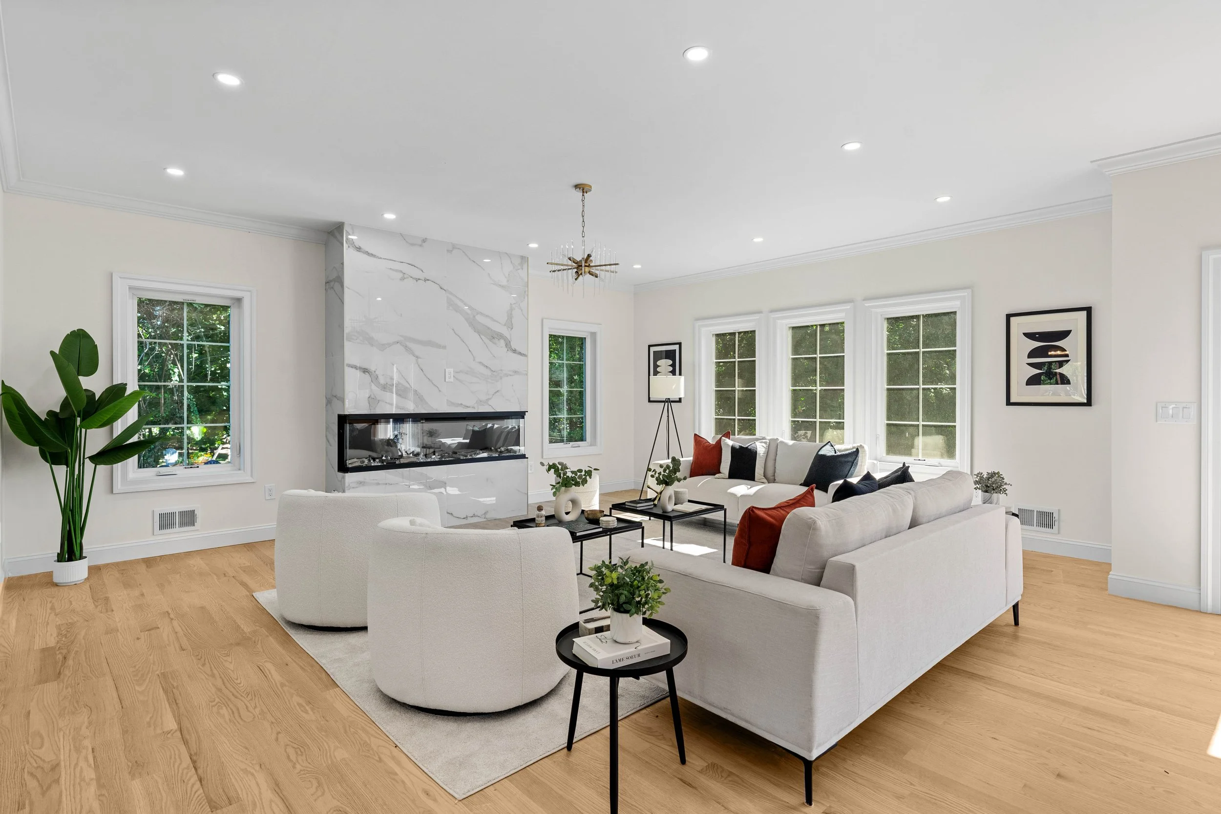 Bright living room with white sofas, armchair, wooden floor, large windows, fireplace with white marble surround, and modern decor.