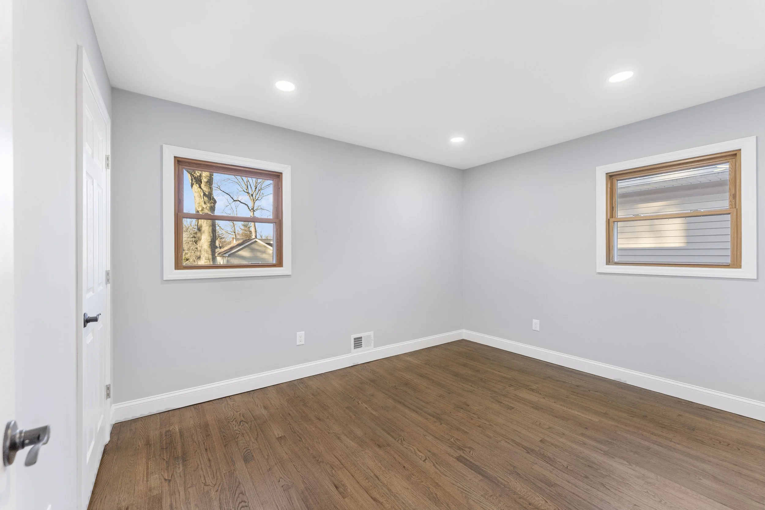 Empty room with hardwood floors, white walls, two windows with wooden trim, ceiling lights, and a white door.