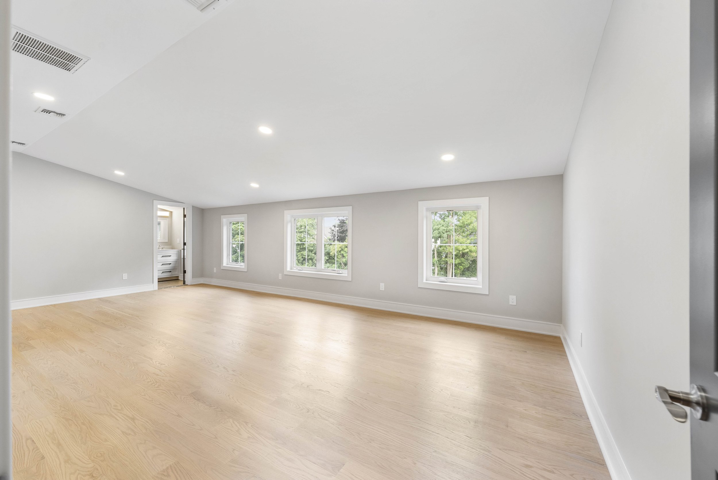 Empty extra room with light wood flooring, white walls, five large windows, recessed ceiling lights, and a small doorway to a bathroom.