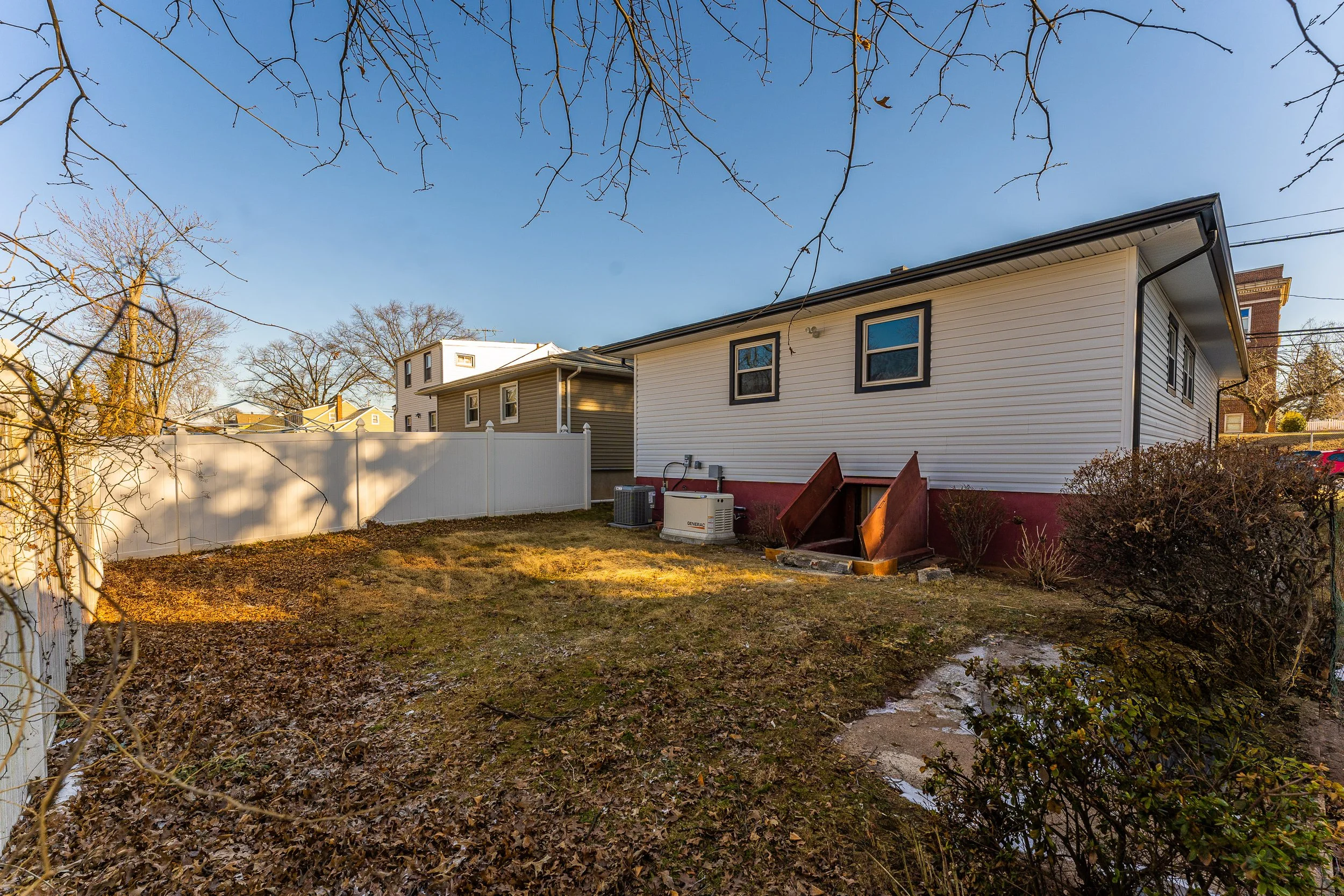 Backyard with patches of snow, leafless trees, and a white fence. House with white siding and small external stairs, air conditioning units, and bushes.