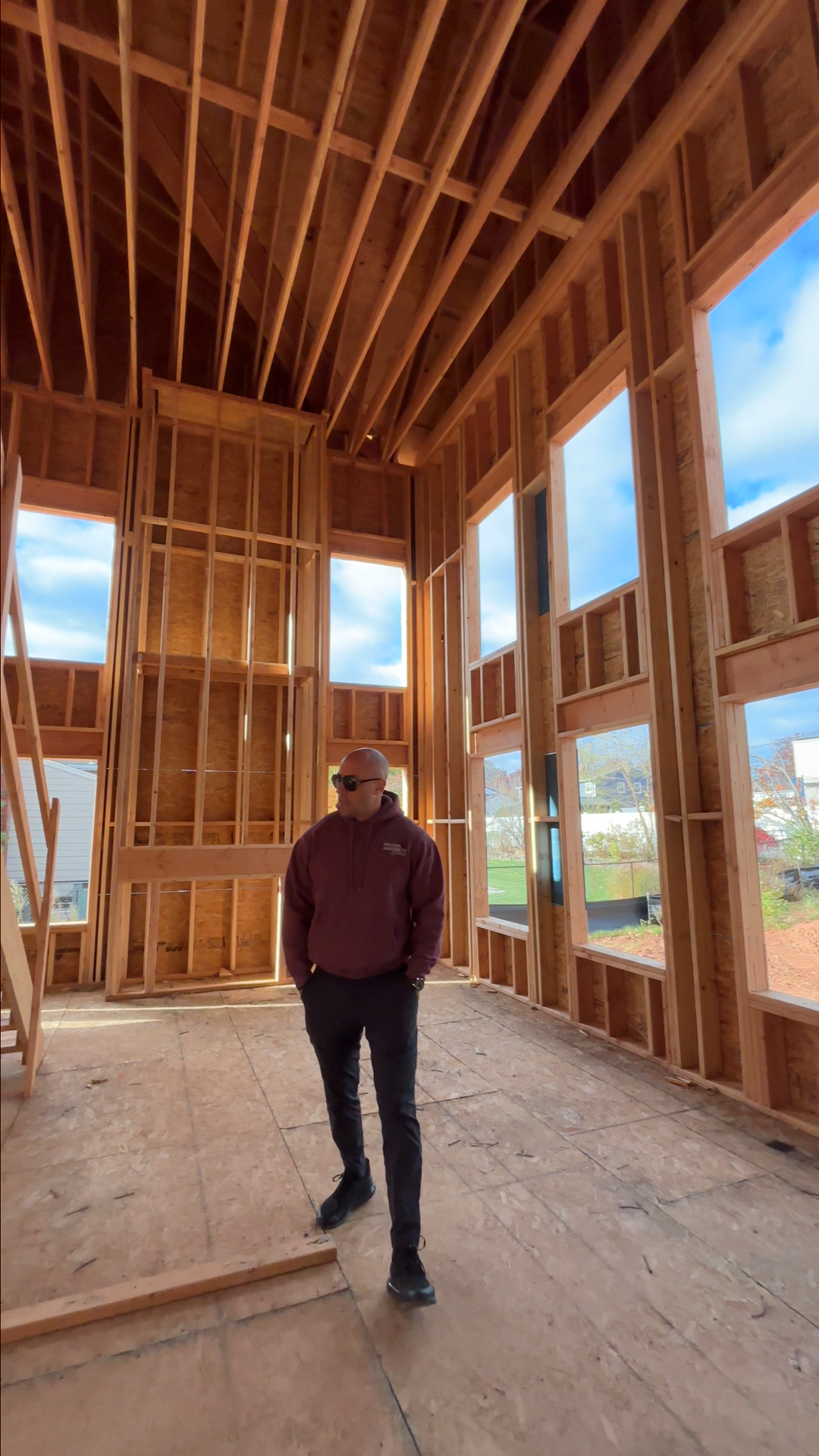 A man wearing sunglasses and a maroon hoodie walking inside the frame of a house under construction with exposed wooden framing and large window openings, overlooking a partly cloudy sky outside.