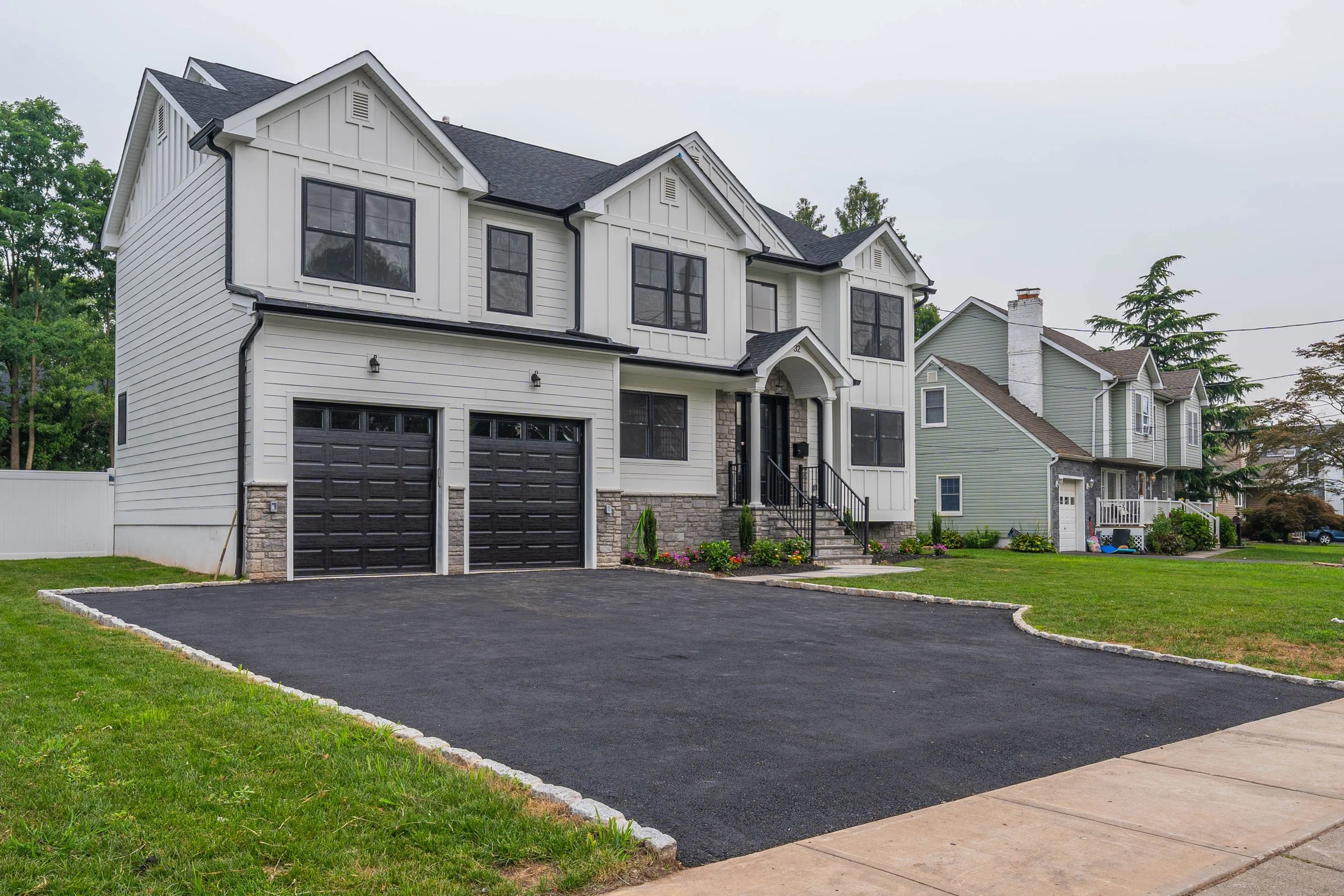 Modern two-story house with a black garage door, front steps, small garden, and green lawn, neighboring houses in background.