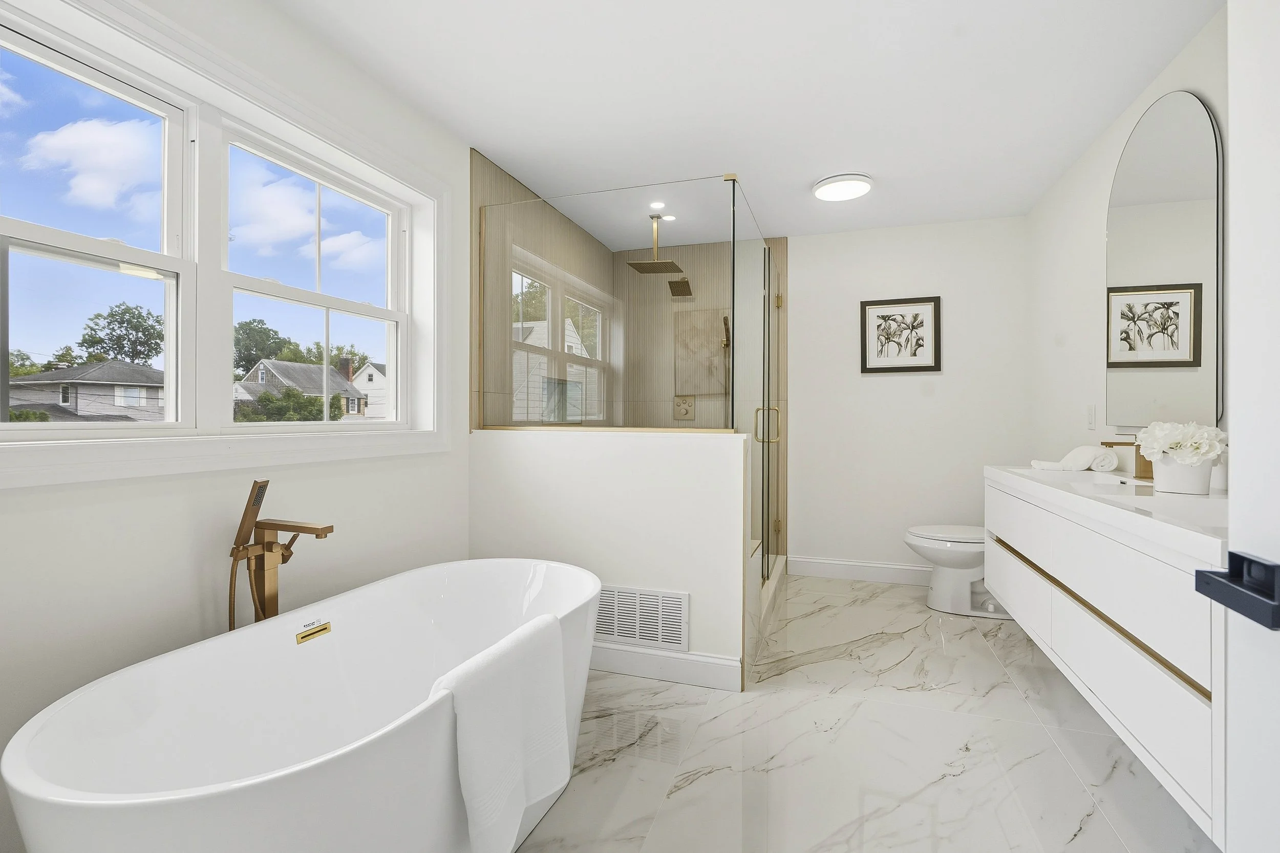 Modern white bathroom with large bathtub, marble flooring, glass shower enclosure, white vanity with mirror, and artwork on walls