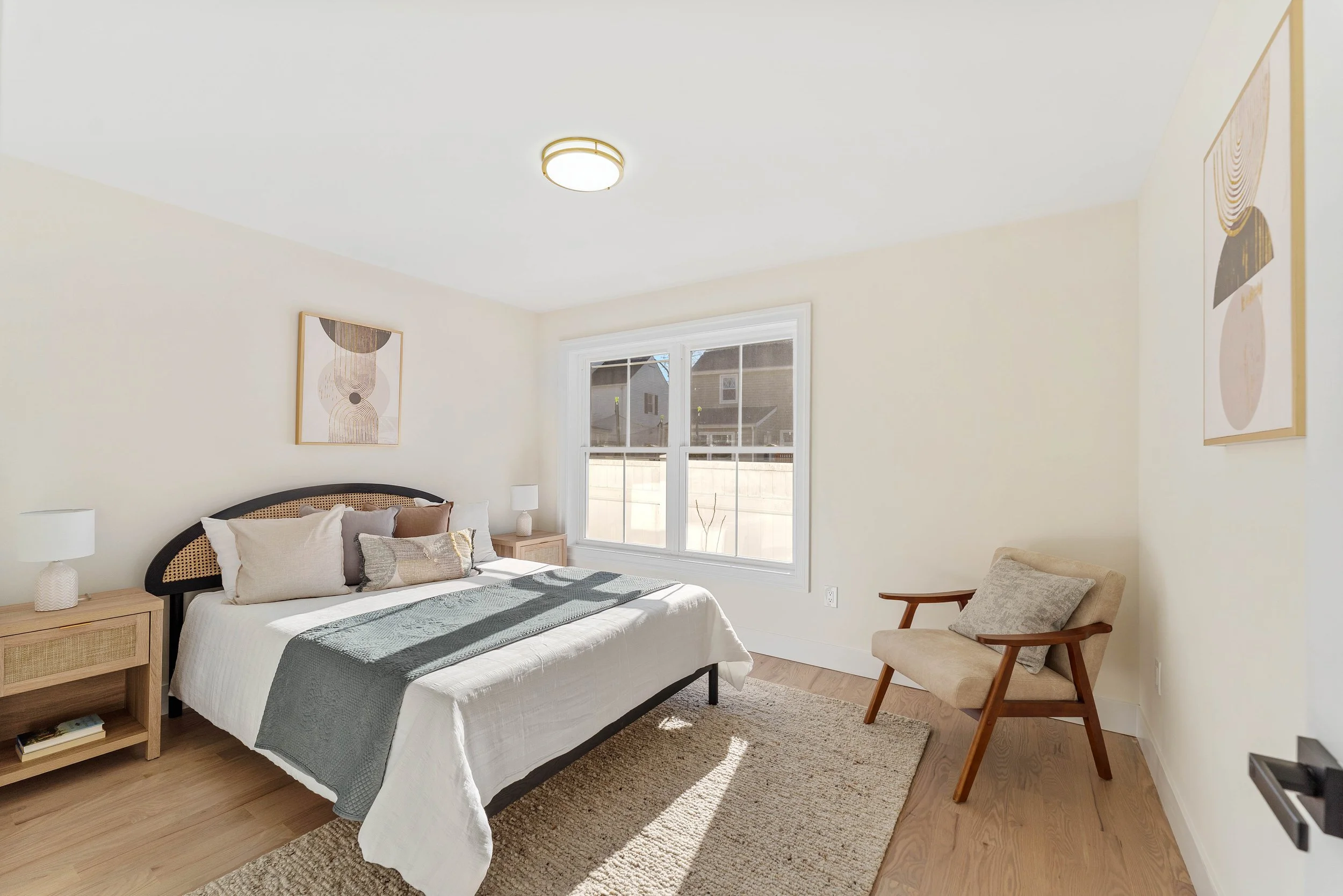 Bedroom with a bed, two nightstands with lamps, a window showing the outside, a beige armchair with a pillow, and modern wall art.