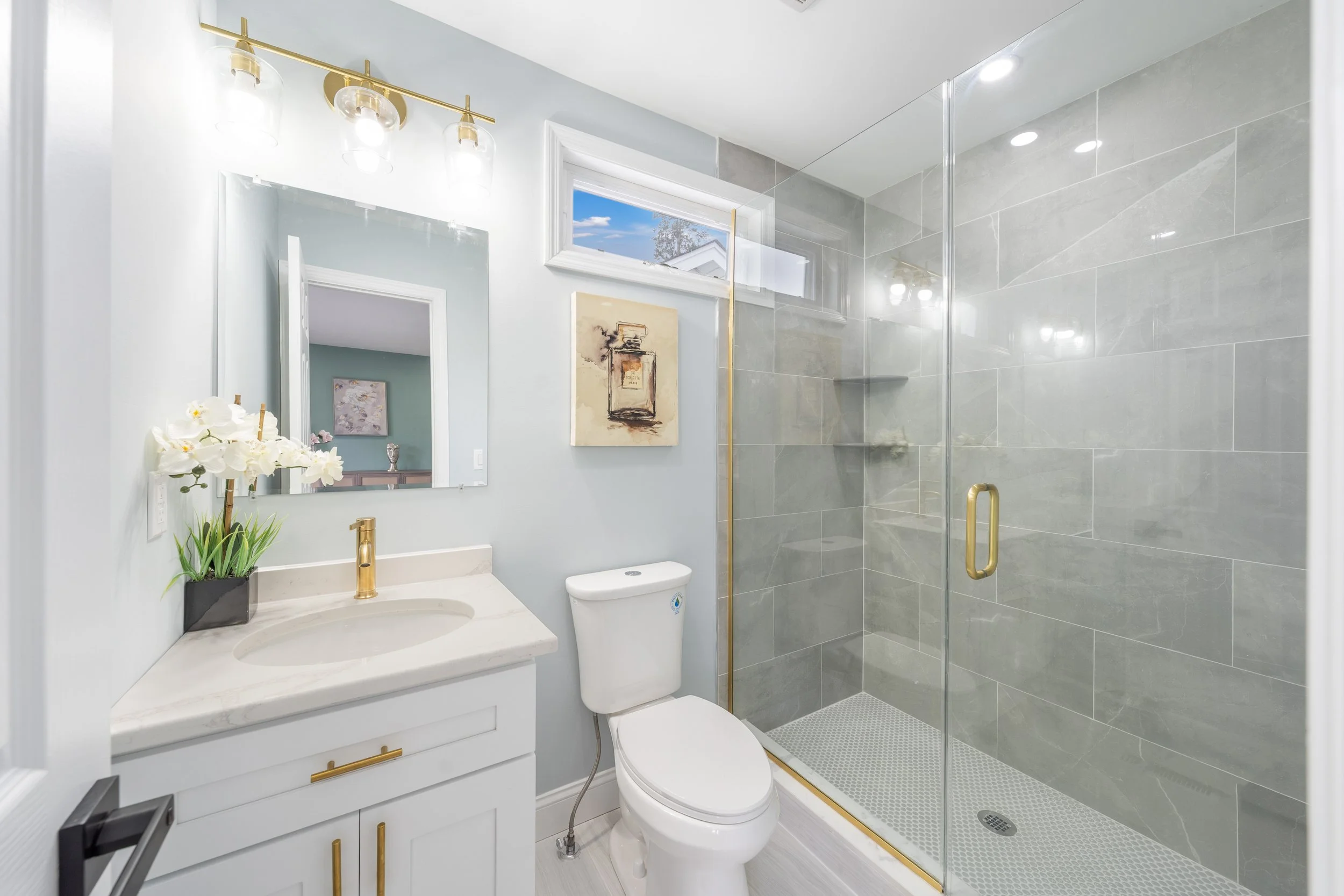 Modern bathroom with white vanity, gold fixtures, a toilet, and a glass-enclosed shower with gray tiles.