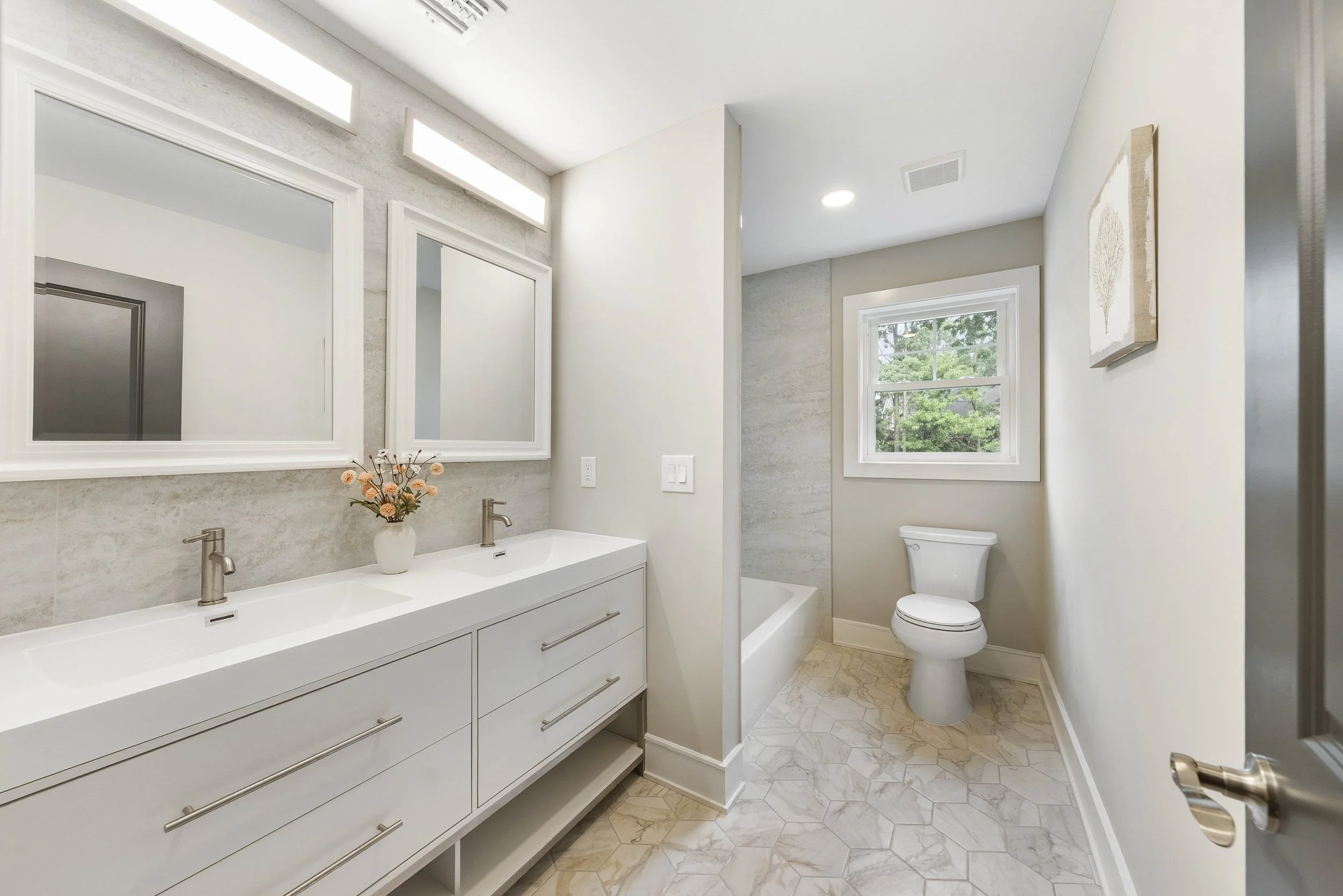 Bathroom with double vanity, two mirrors, a flower vase, bathtub, toilet, window, and framed artwork.