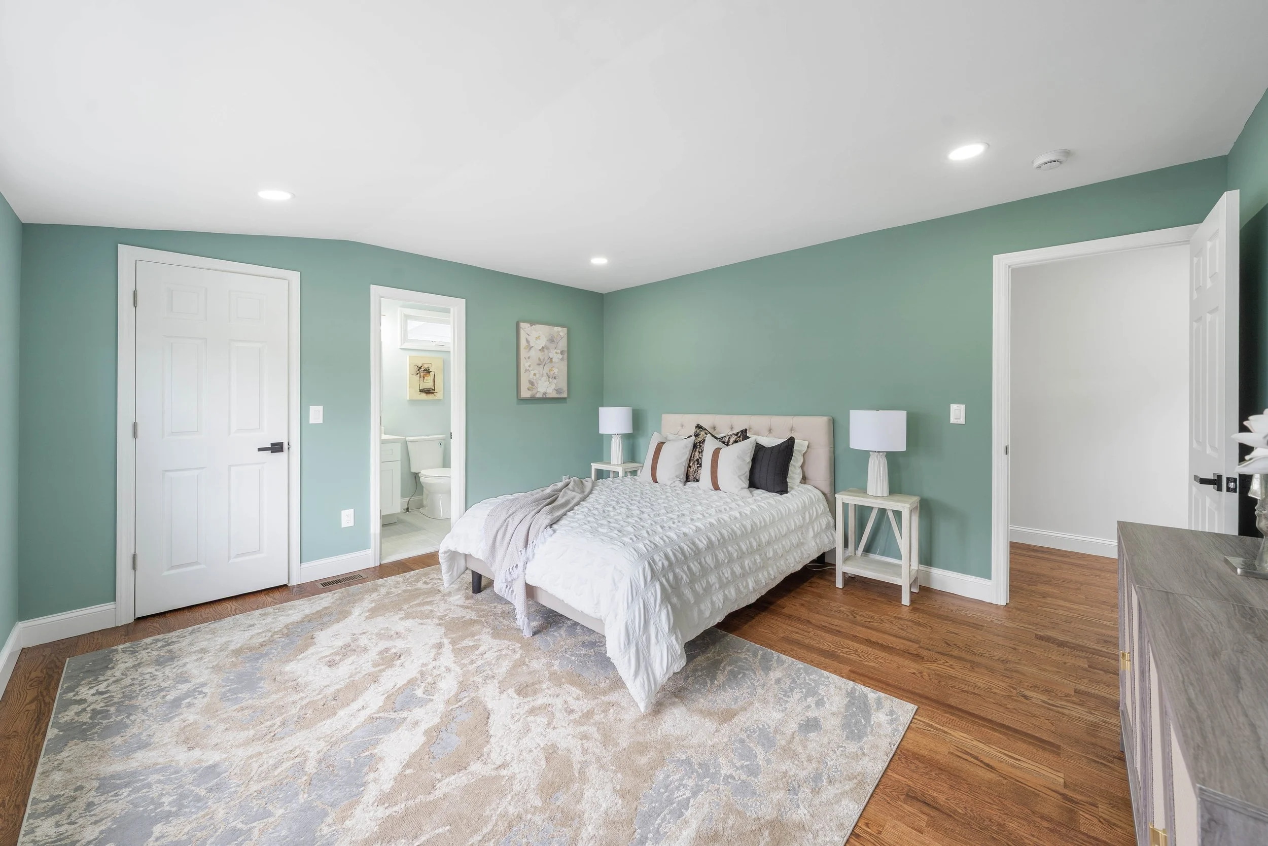 A bedroom with a bed, two nightstands with lamps, a dresser, artwork on the wall, and a bathroom visible through an open door.