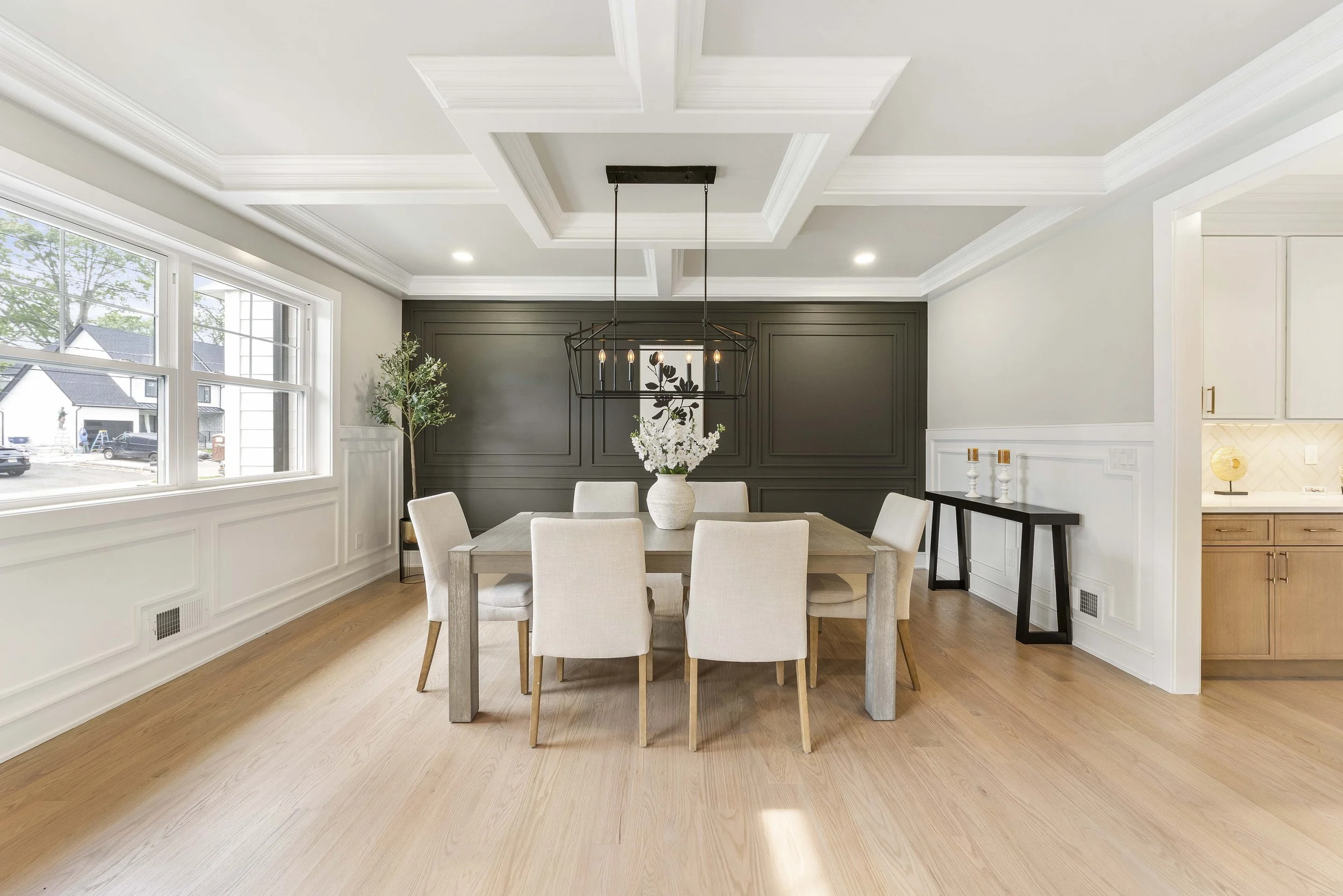 Bright dining room with eight beige chairs around a wooden table, a large vase with white flowers, large windows, a black wall panel, and a chandelier hanging from a ceiling with decorative molding.