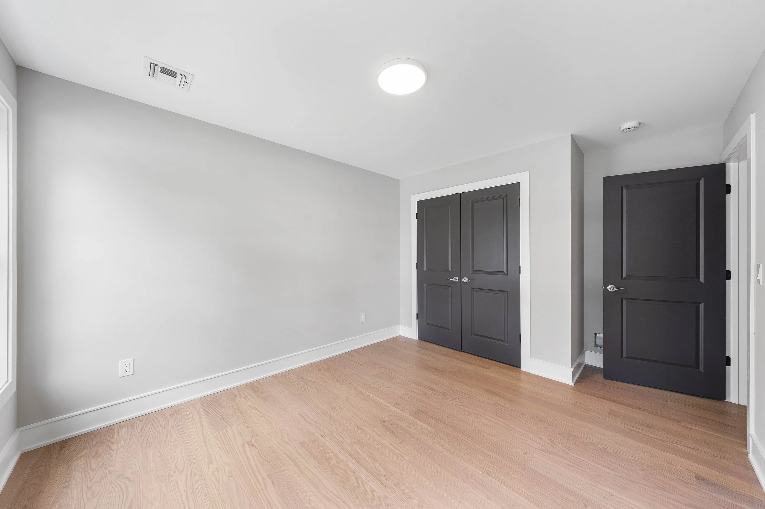 Empty room with light wood floors, white walls, a white ceiling with a round light, black double doors, and an open black door to a hallway.