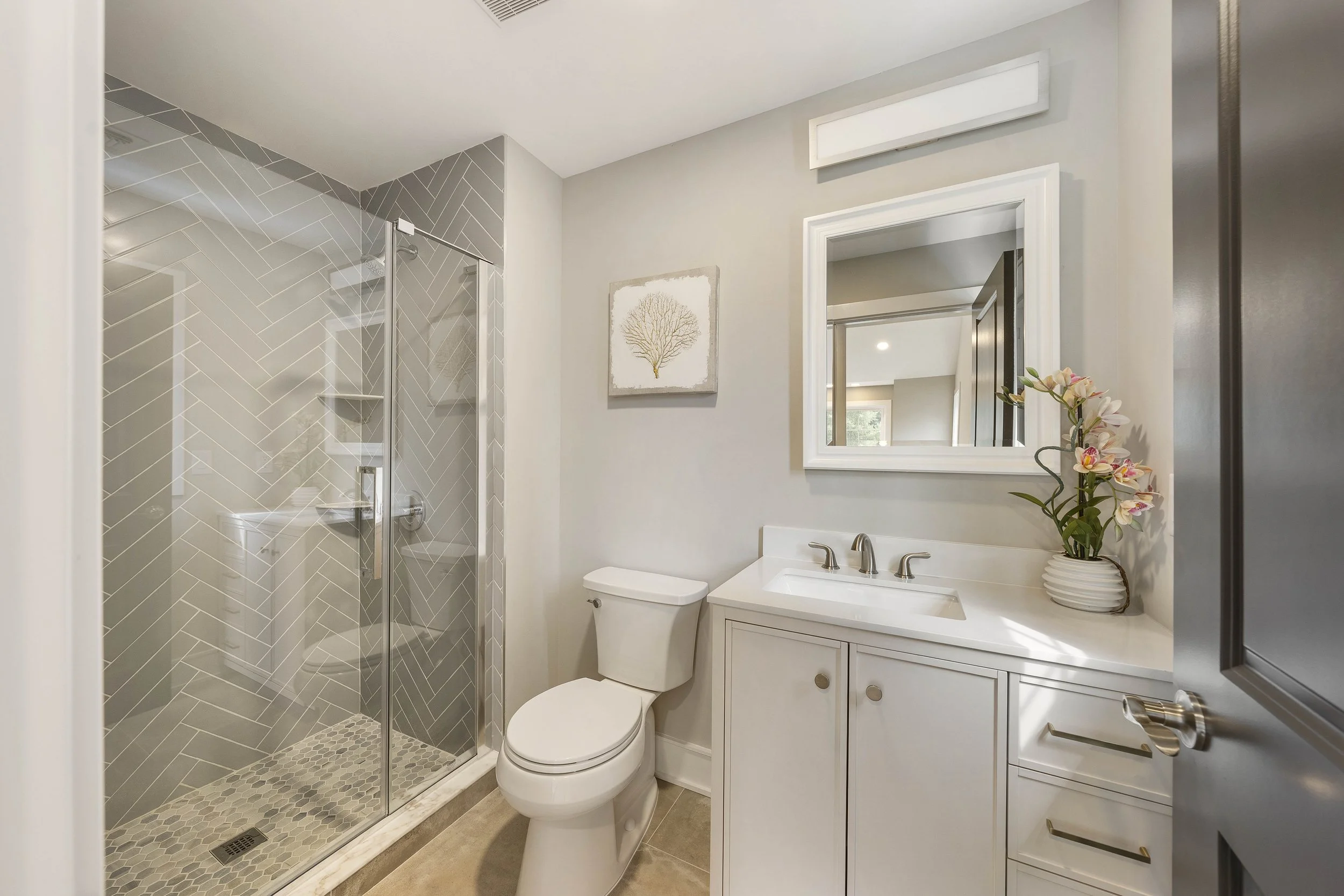 Modern bathroom with glass-enclosed shower, white toilet, white vanity with a sink, mirror, and decorative orchids, light-colored walls, and artwork.