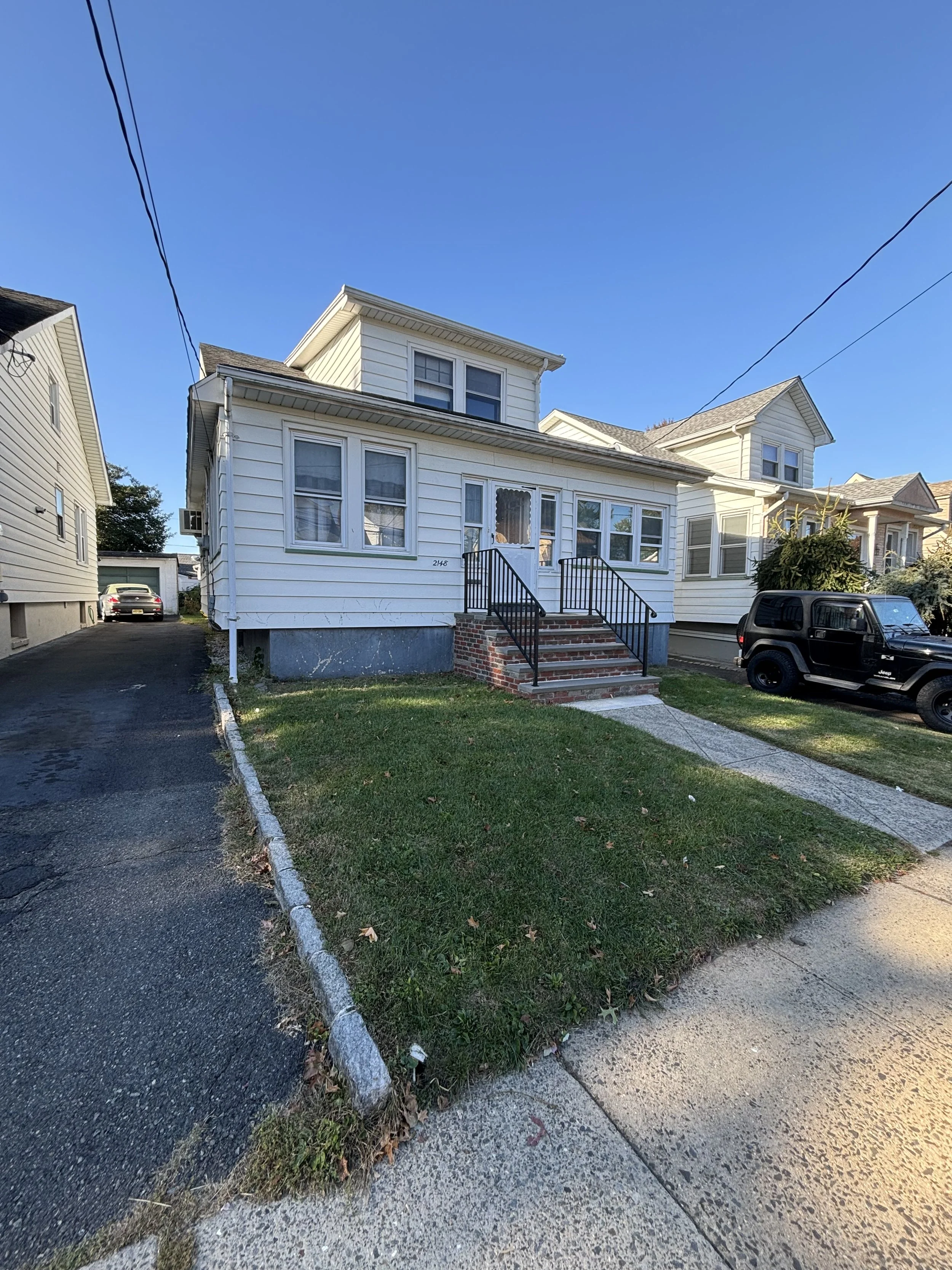 White house with brick steps and black handrails, small front lawn, driveway on the left, and black SUV parked on the right, under a clear blue sky.
