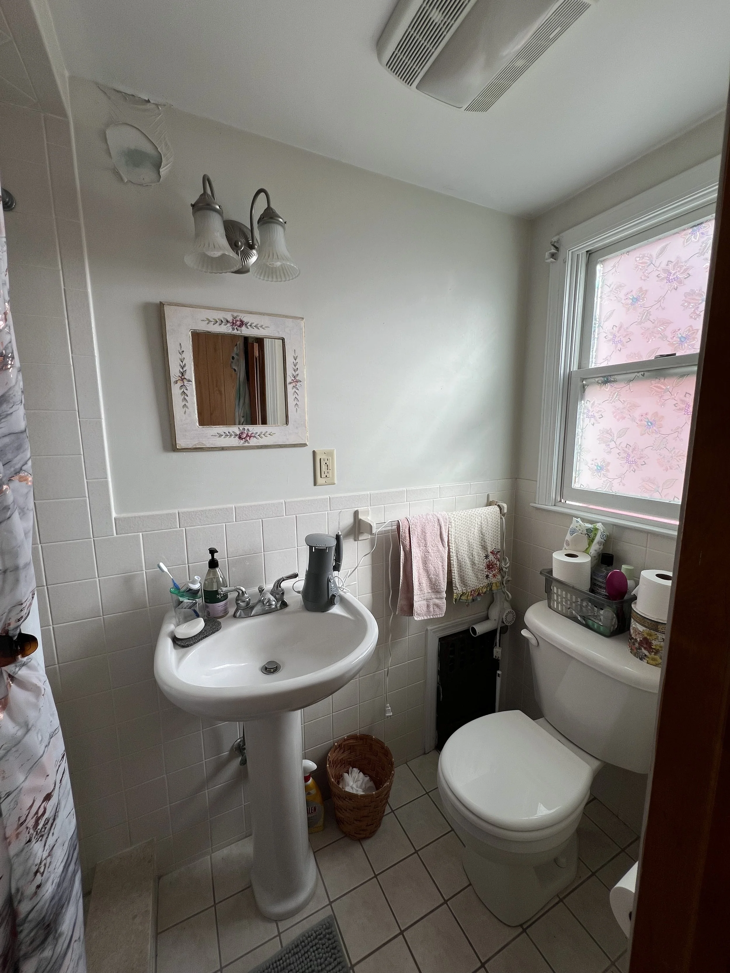 Small bathroom with a white pedestal sink, a mirror with a floral frame, a toilet with a basket and toiletries on top, a window with floral blinds, and a towel rack, with a wall-mounted light fixture and an air vent on the ceiling.