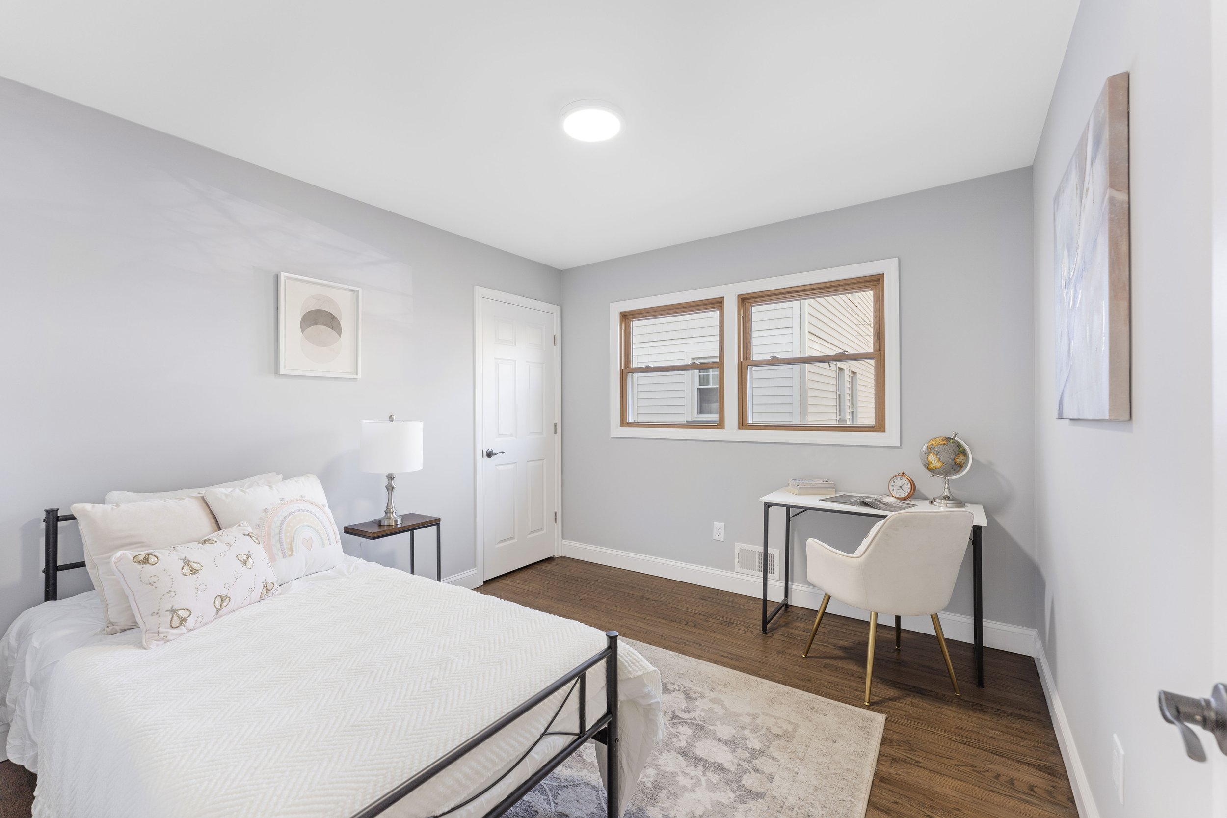 A bedroom with light gray walls, a bed with white bedding and decorative pillows featuring rainbows and butterflies, a wooden nightstand with a table lamp, a desk with a globe, book, and clock, and a white armchair.