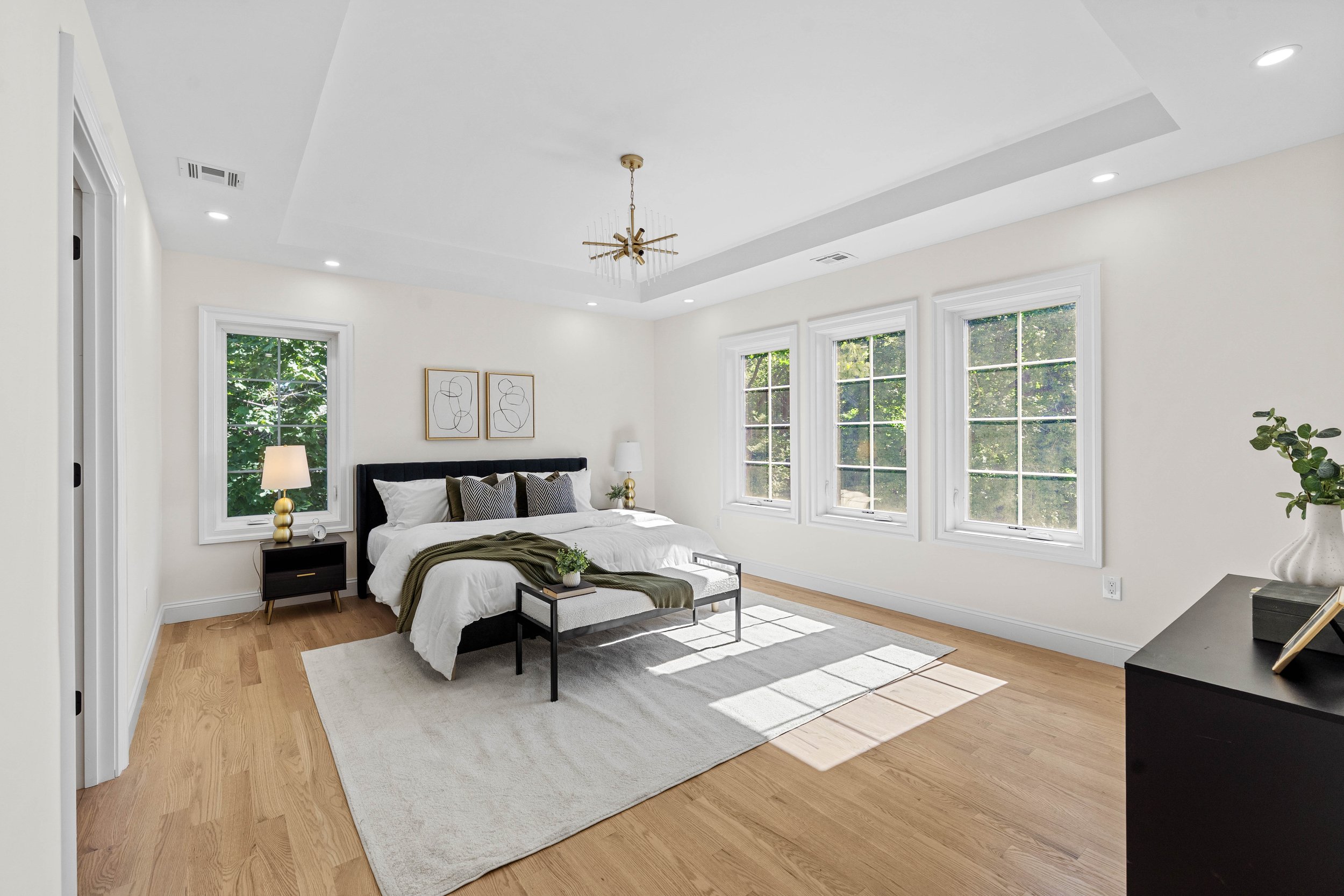 Bright bedroom with a bed, nightstands, lamps, and large windows showing greenery outside.