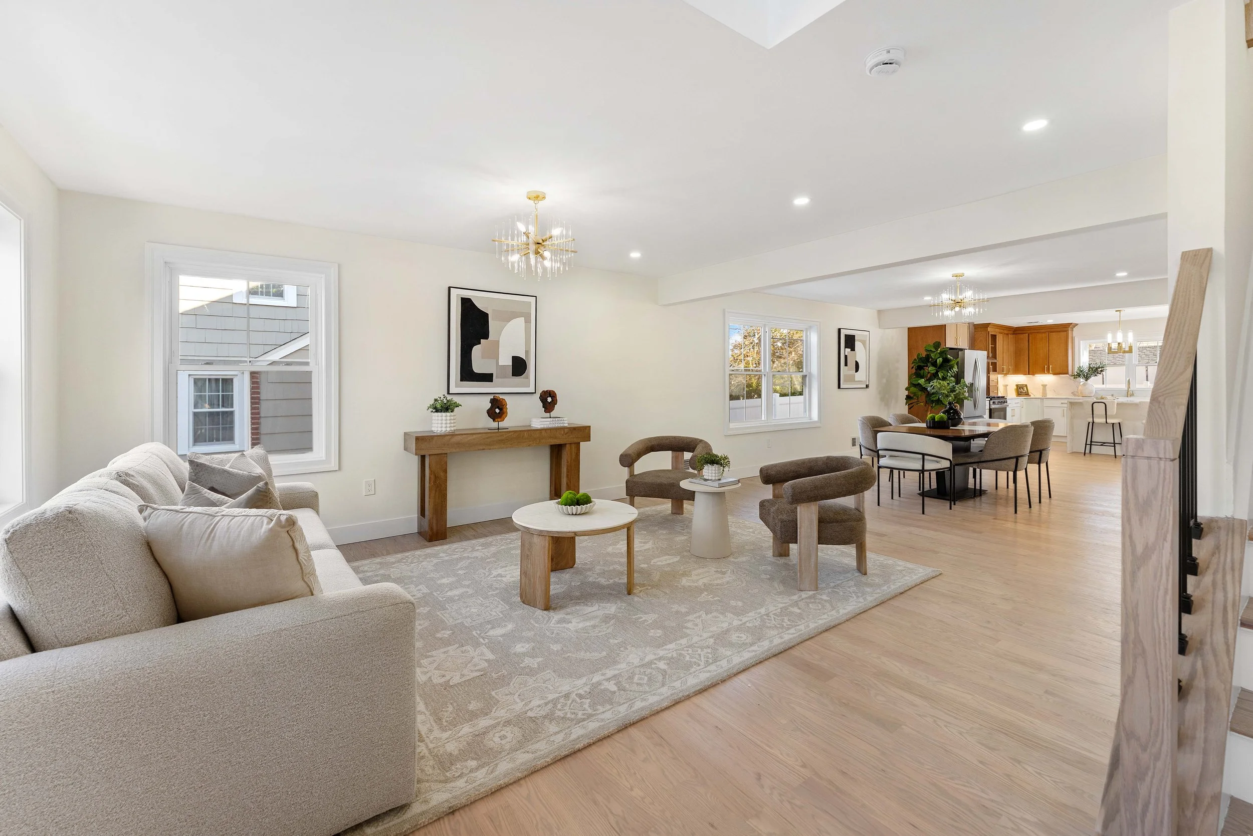 Open-concept living and dining area with beige sofa, wooden tables, modern artwork, and a white kitchen with wooden cabinets in the background.
