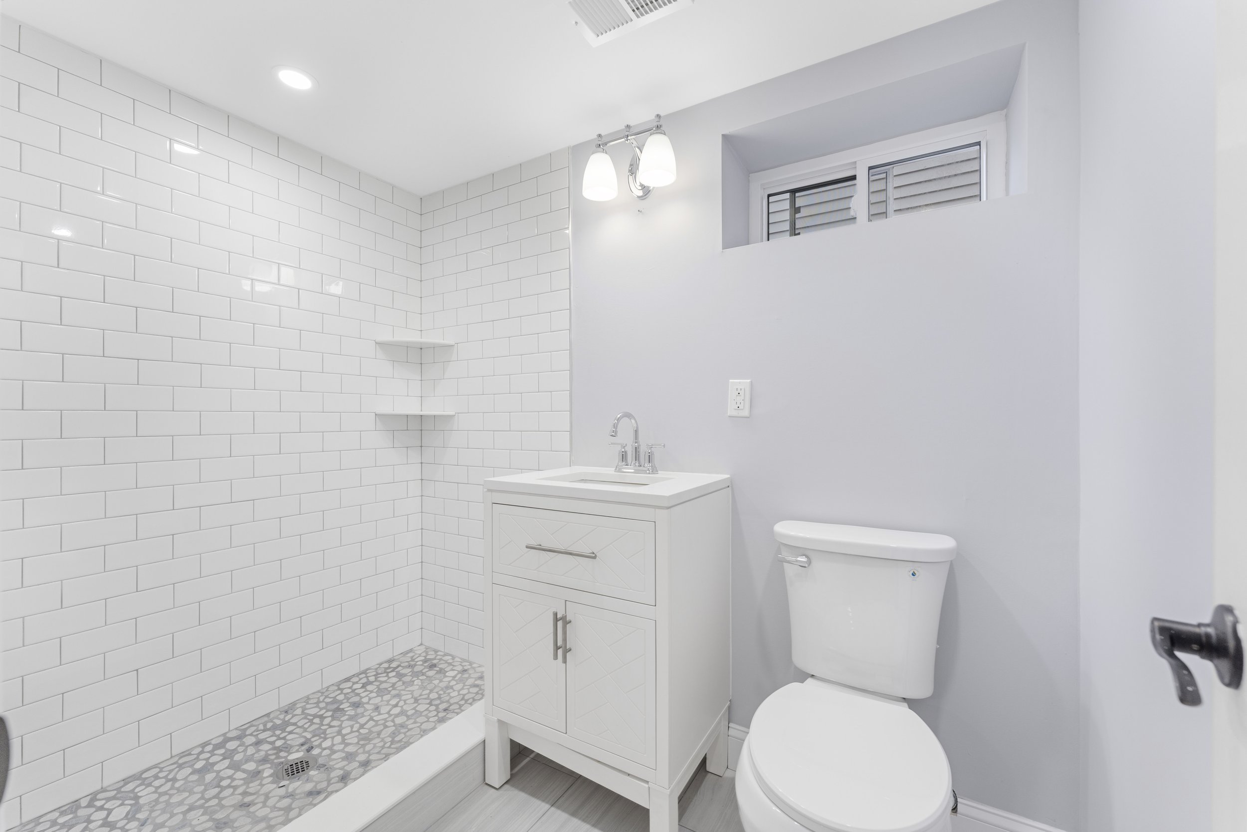Small bathroom with white tiled walk-in shower, white vanity with sink, and toilet. Light gray walls and a small horizontal window above the toilet. Recessed ceiling light and wall-mounted light above the mirror.