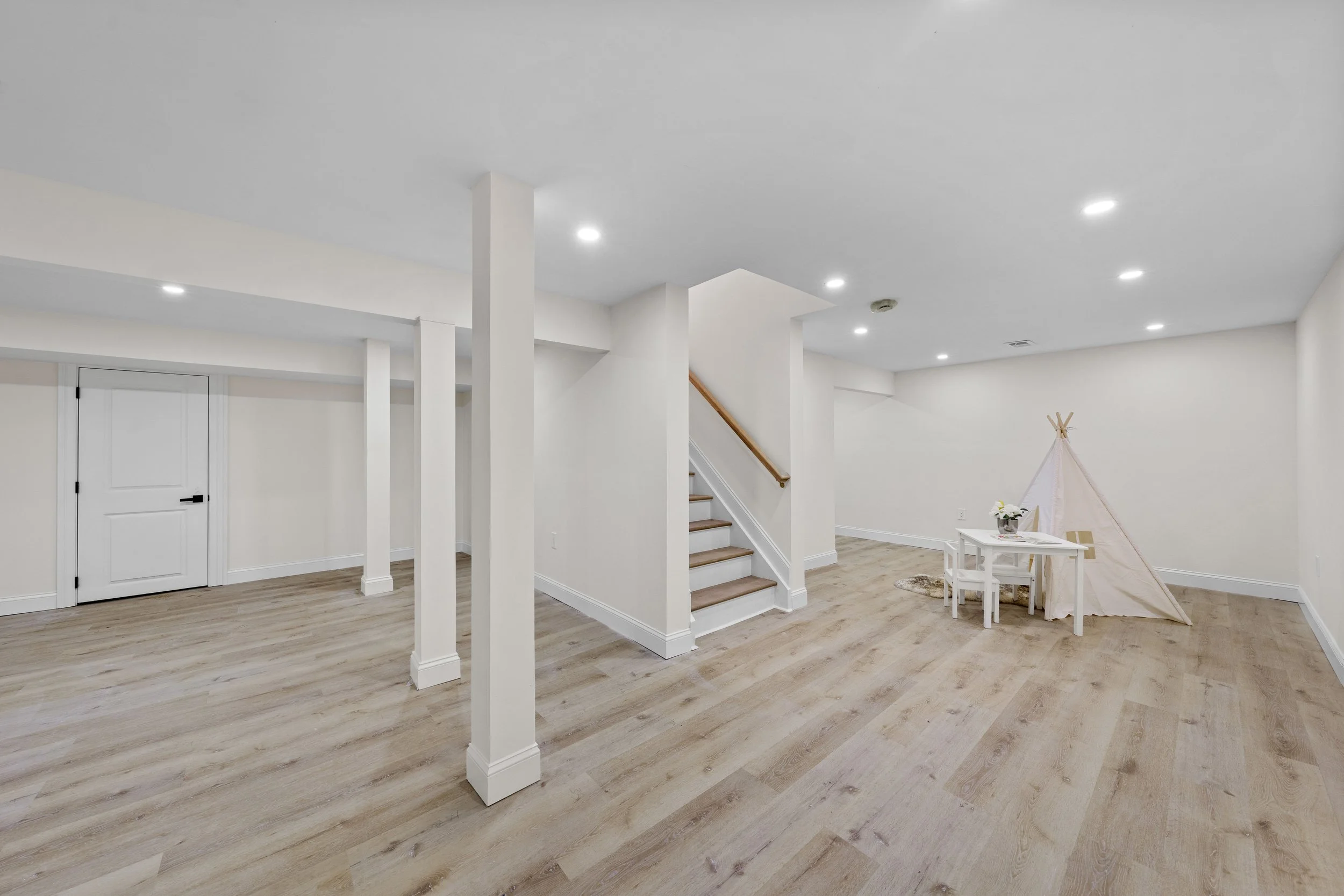Empty room with light wood flooring, white walls, ceiling lights, a small white table with two chairs, a small teepee tent, and a staircase with wooden handrail.