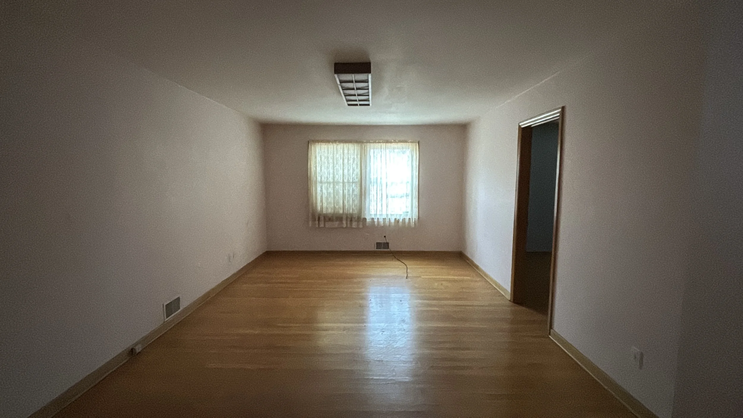 Empty living room with wooden floors, a window with lace curtains, and a doorway on the right.