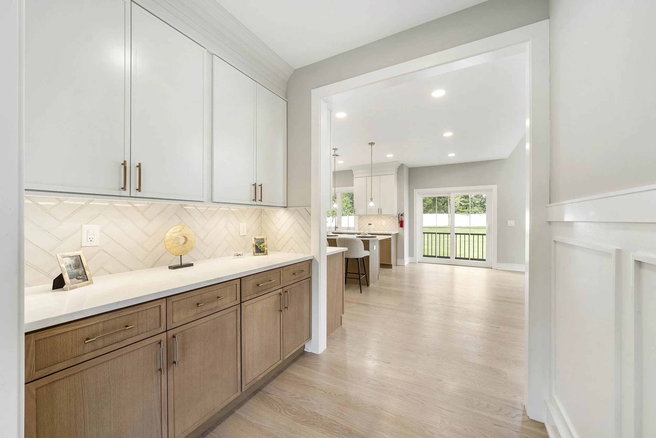 Modern open-plan kitchen with white upper cabinets and wooden lower cabinets, light wood flooring, and large sliding glass door leading to a backyard.