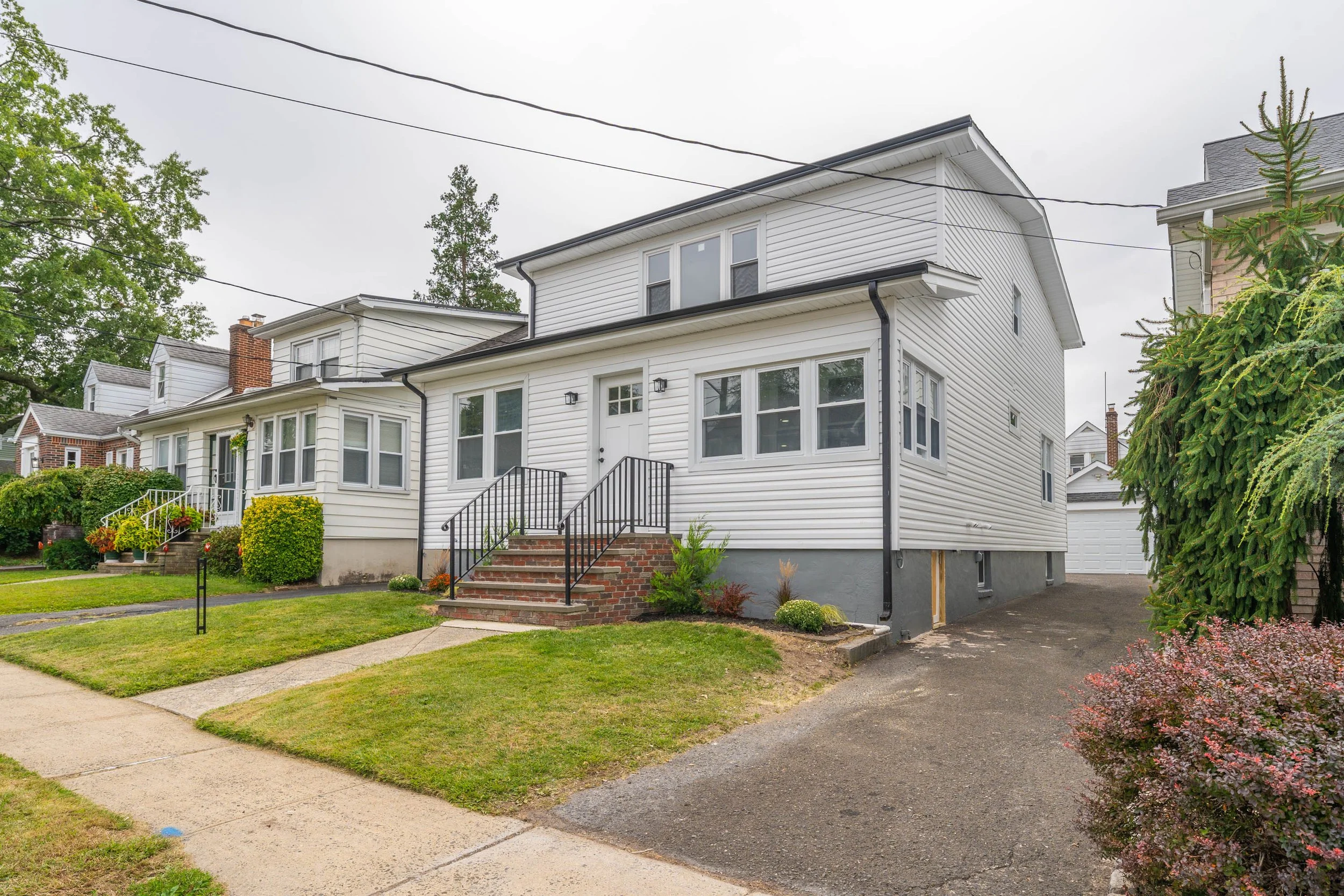 A white, two-story house with a small front porch and brick stairs. The house has multiple windows and a gray foundation. The yard is landscaped with bushes and a small lawn, and there's a driveway on the right side leading to a detached garage. Over