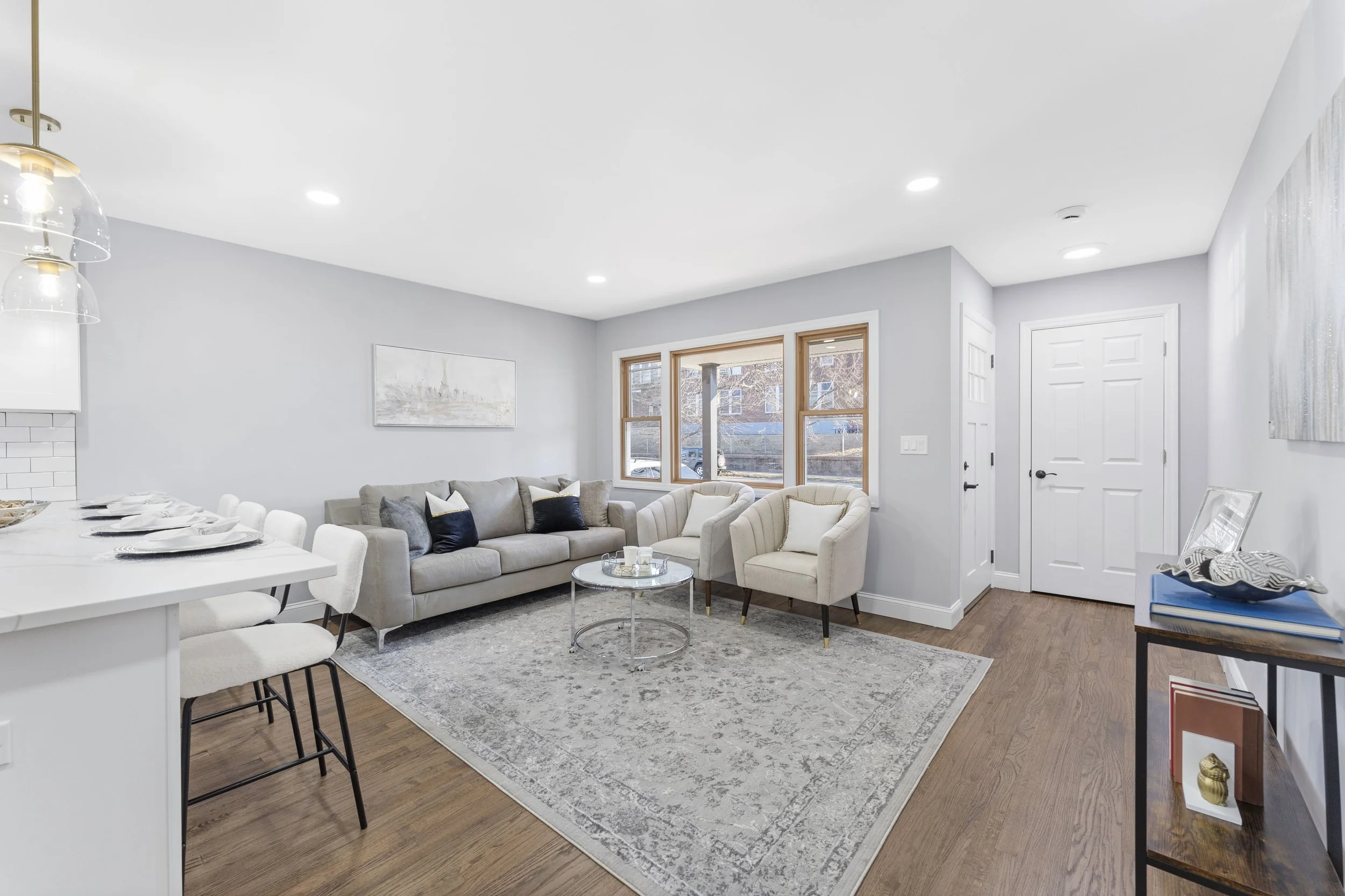 Bright living room with gray walls, white ceiling, hardwood floors, and large windows, furnished with a gray sofa, two white armchairs, a round glass coffee table, and a patterned area rug.