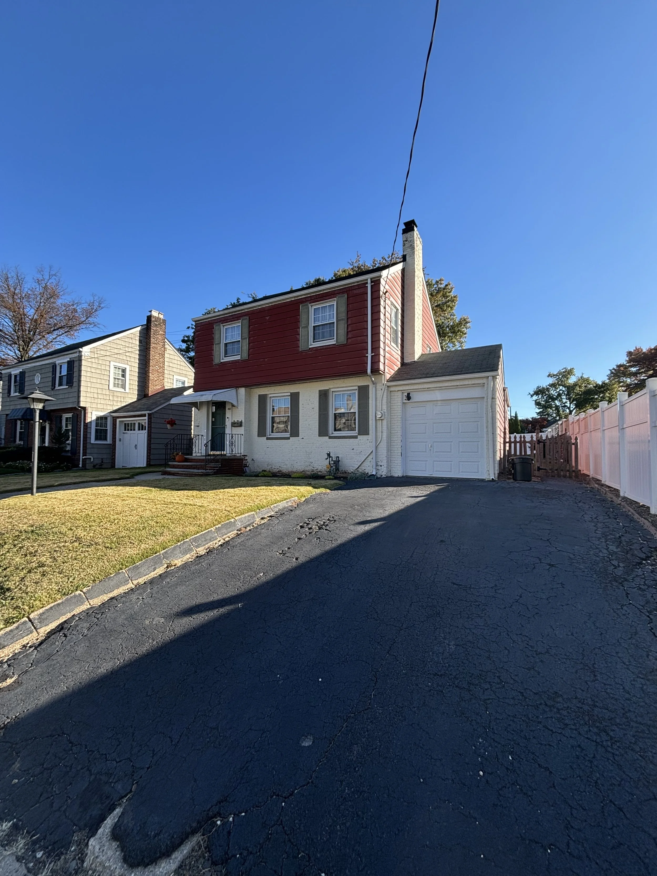 A residential house with a two-story structure, red upper siding, white lower walls, a white garage door, and a driveway. The house has windows with light-colored shutters and a small front lawn with a lamppost. The sky is clear and blue.