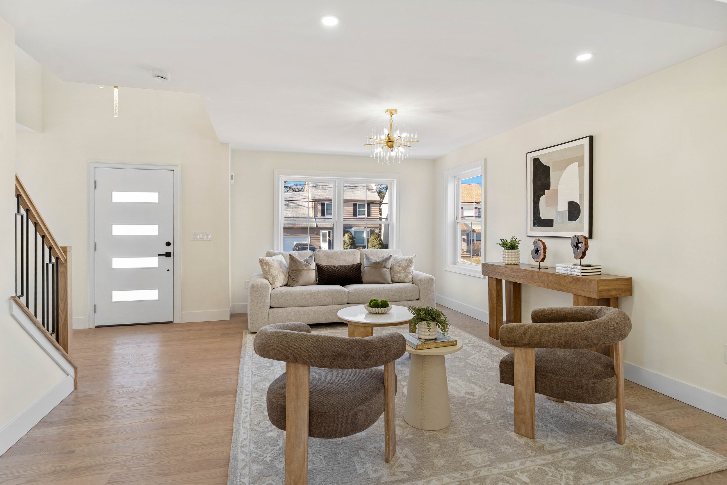Living room with beige sofa, two brown accent chairs, light wood coffee table, side table, and wall art, with large windows and hardwood floors.