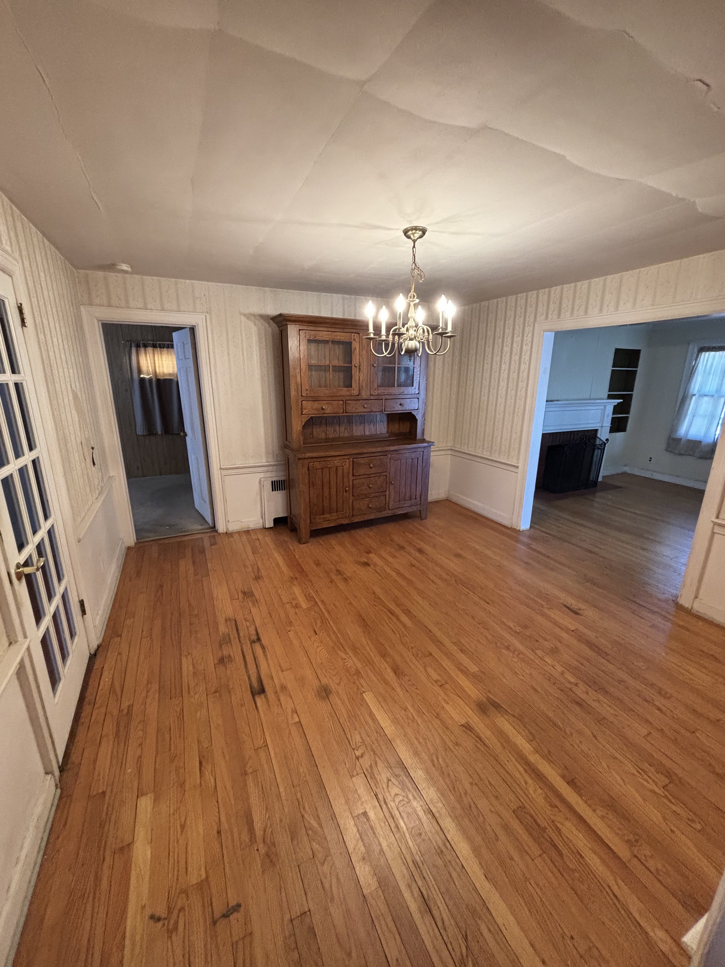 Empty dining room with hardwood floors, a wooden china cabinet, a chandelier, and a doorway to another room with a fireplace and window.