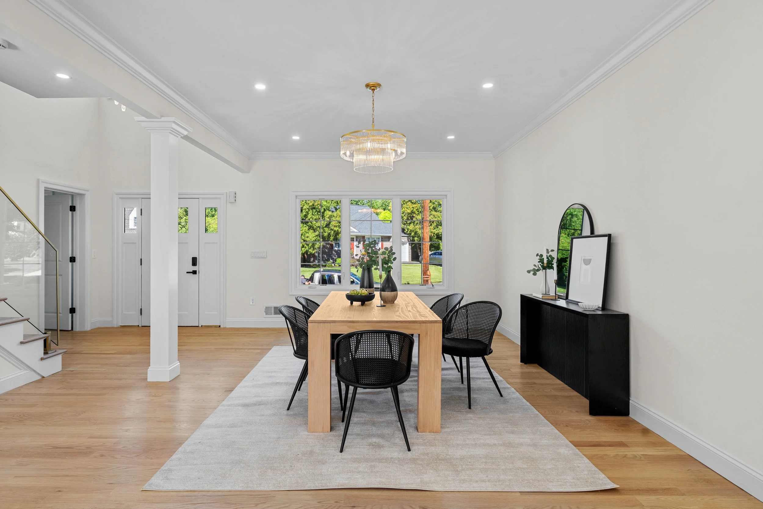 Dining room with a wooden table, six black chairs, a black sideboard with a mirror and plants, a chandelier, and a window showing a street view.