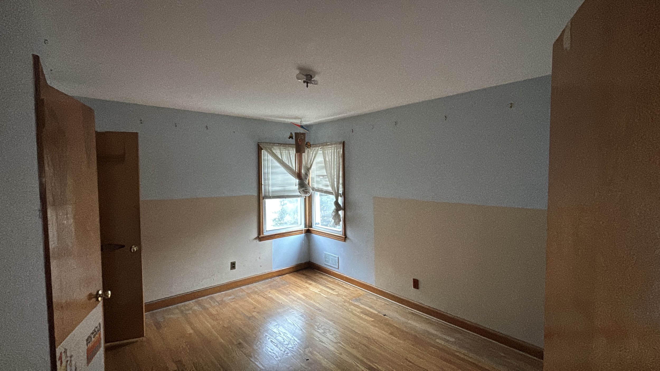 Empty room with hardwood floors, light blue and beige walls, a window with closed blinds and torn curtains, and an open wooden door.