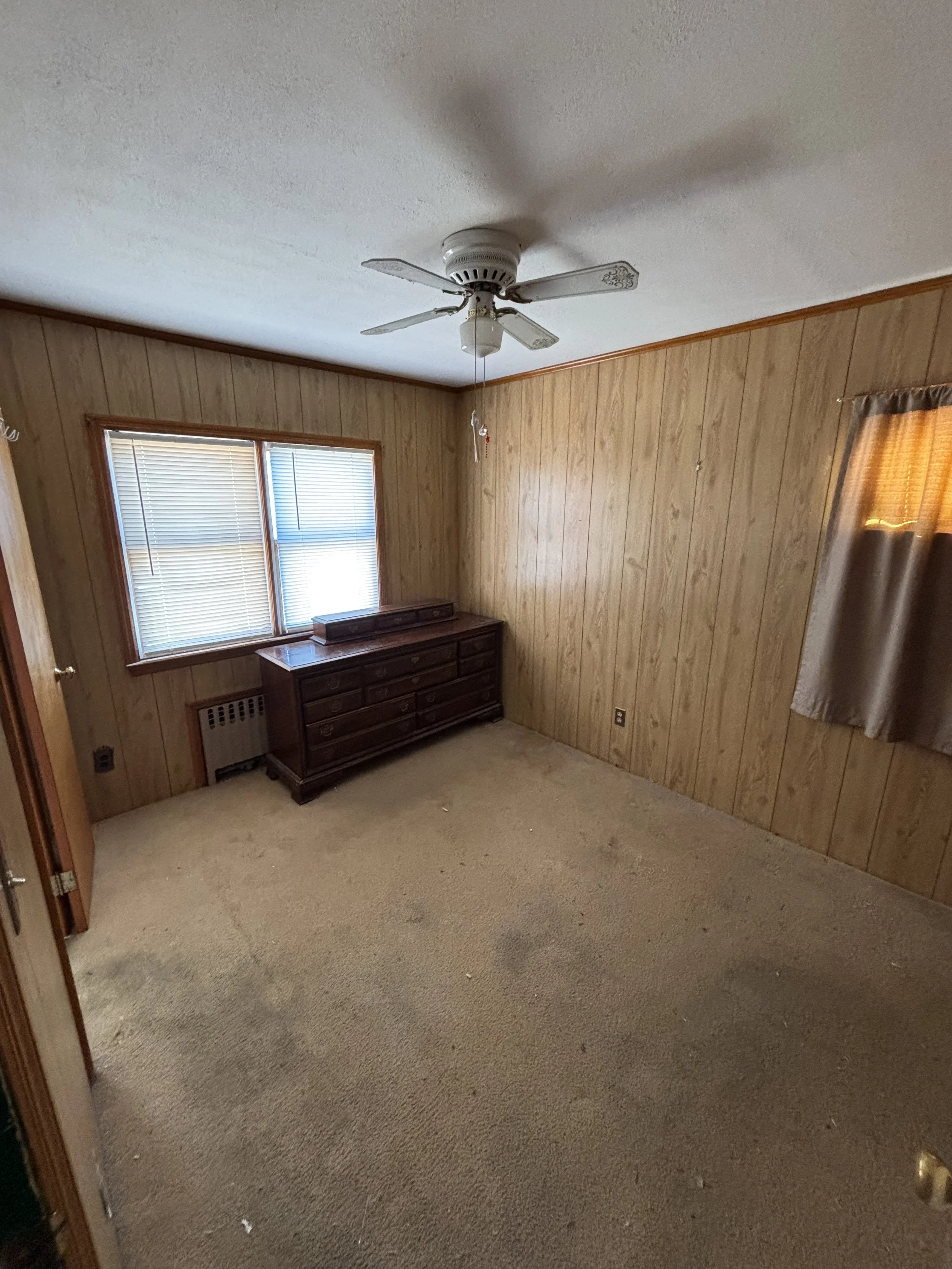 Empty room with wood paneling, a ceiling fan, window with blinds, and a wooden dresser.