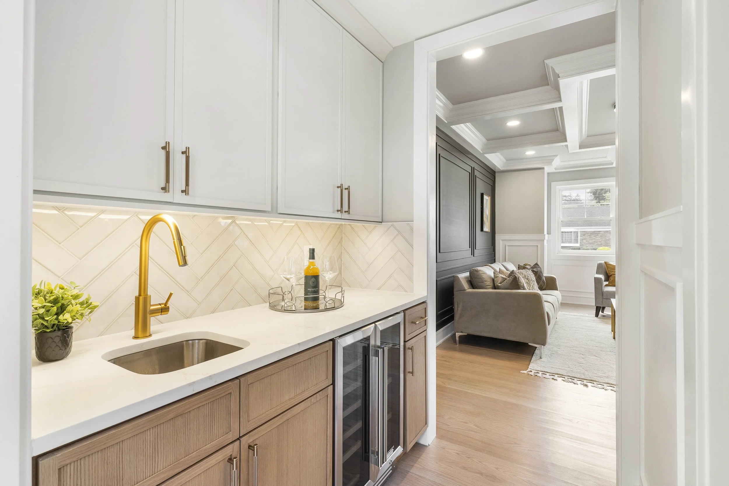 A view from the kitchen into the living room, showing a kitchen countertop with a small sink, a gold faucet, a potted plant, a bottle of wine, and glasses, with cabinets above and a mini fridge below. The living room has a gray sofa, a window, and a 